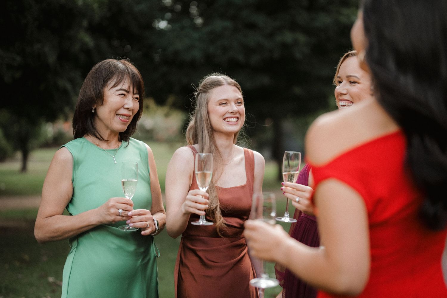 Women in elegant dresses laughing and holding champagne glasses at an outdoor garden party celebration.
