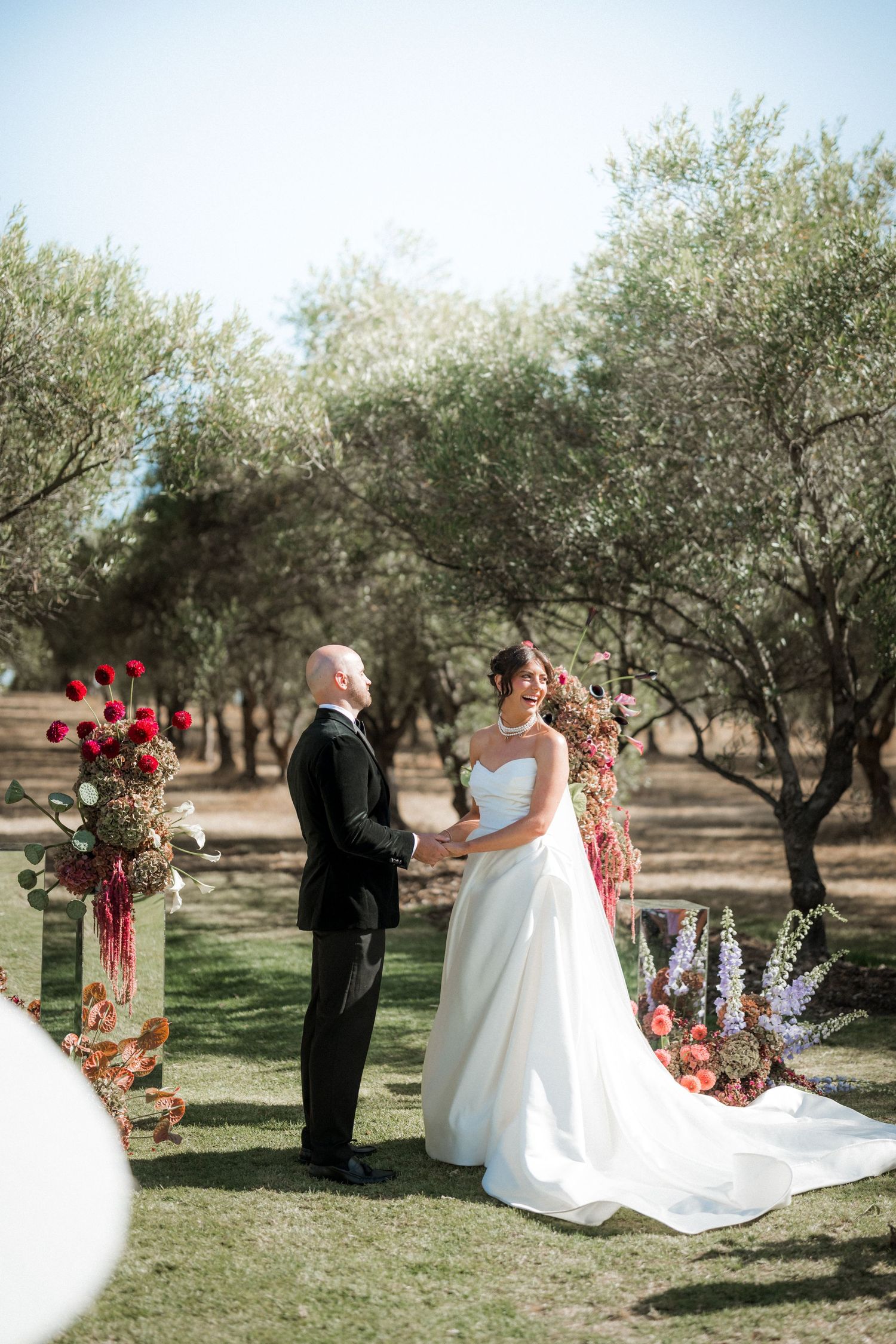 Outdoor wedding ceremony takes place in a serene olive grove with floral arch decorations.