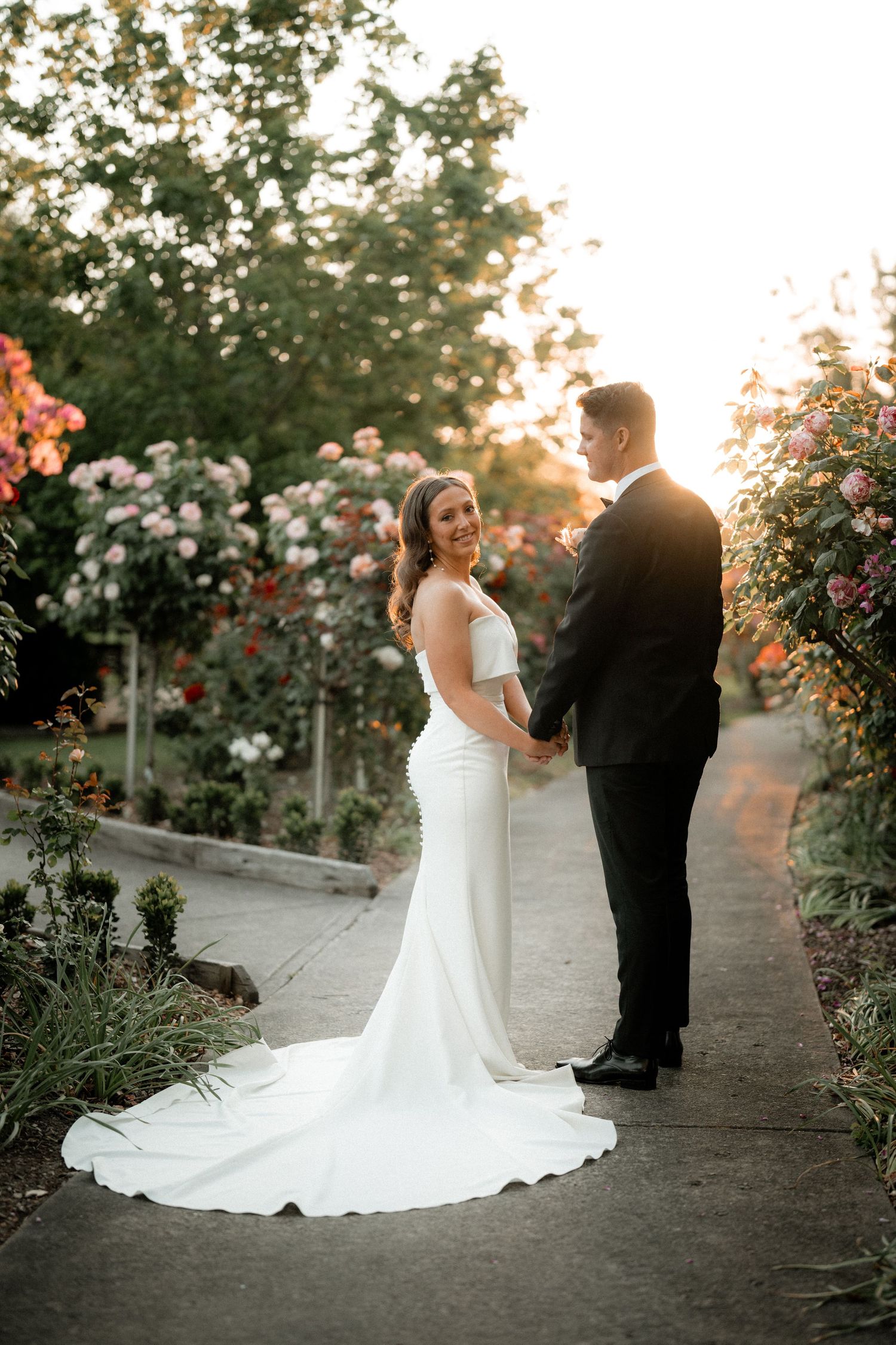 Newlywed couple poses along garden path with blooming roses at sunset in romantic wedding photography series.