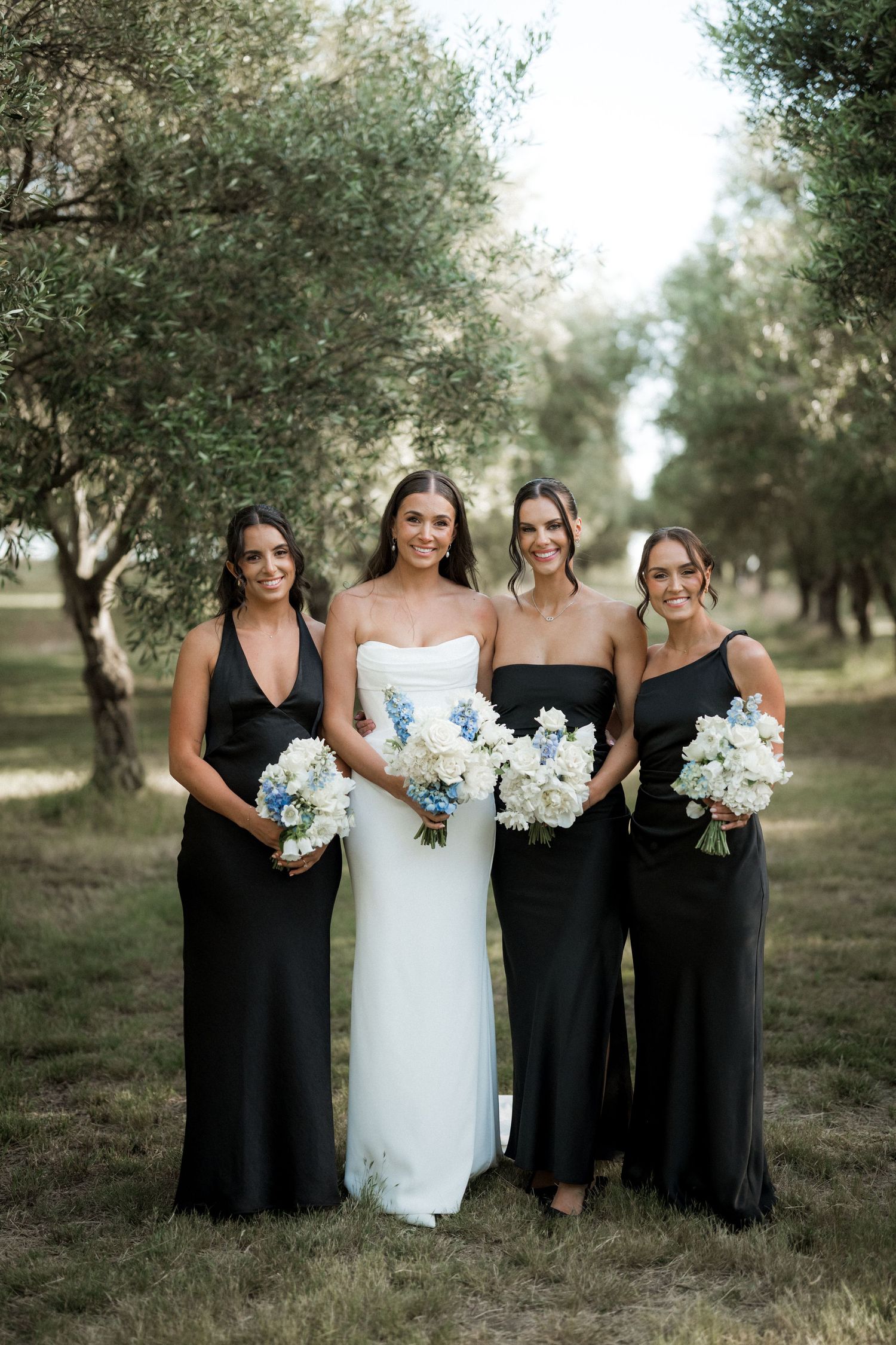 A bridal party in black dresses and white bouquets poses together in an olive grove setting.