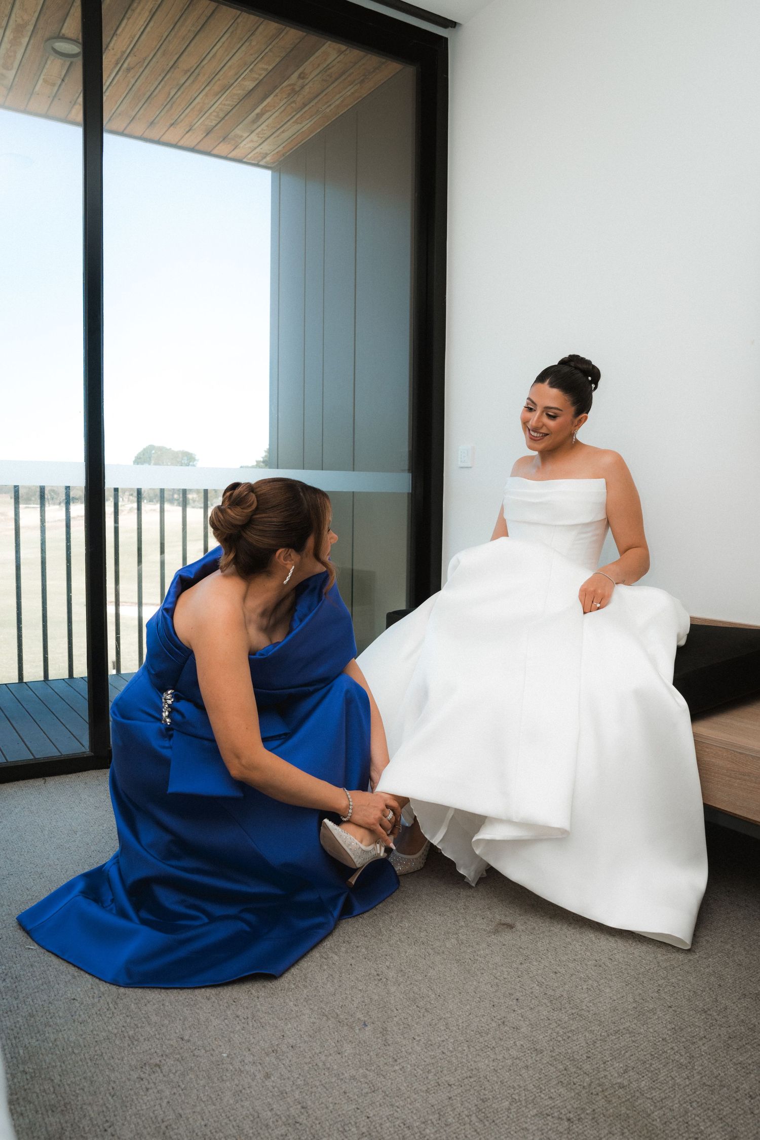 Bride in royal blue bridesmaid dress sits on grey carpet next to flowing white wedding gown.