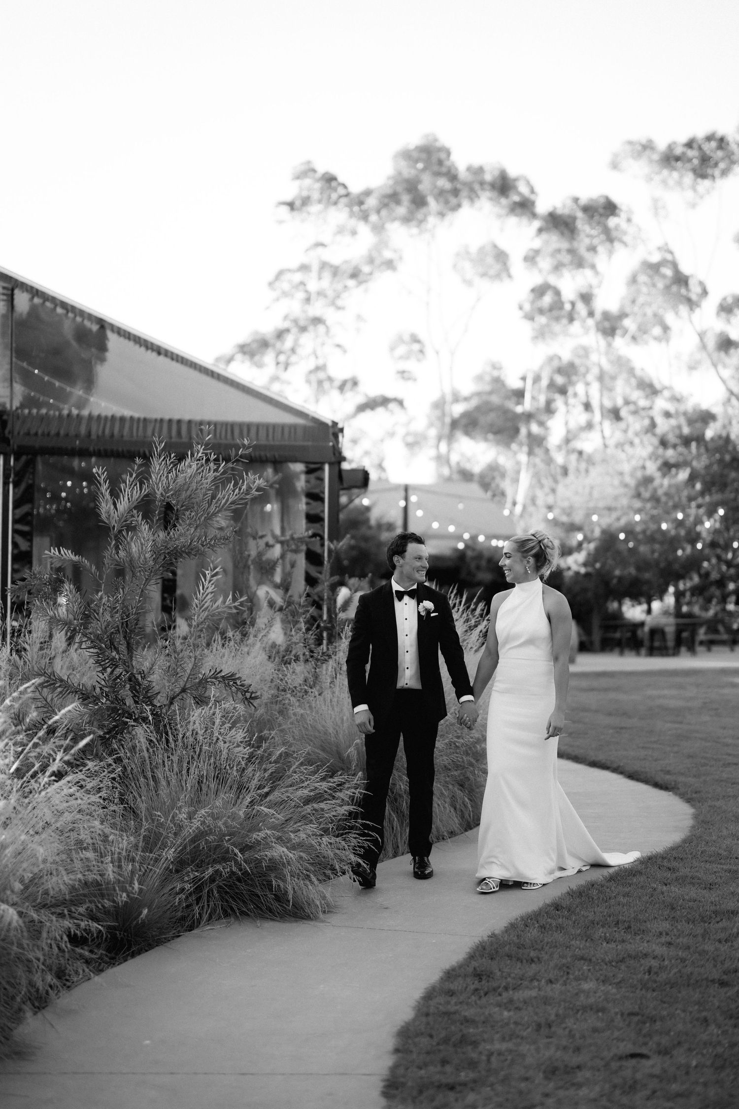 A couple walks along a garden pathway in formal wedding attire during golden hour.