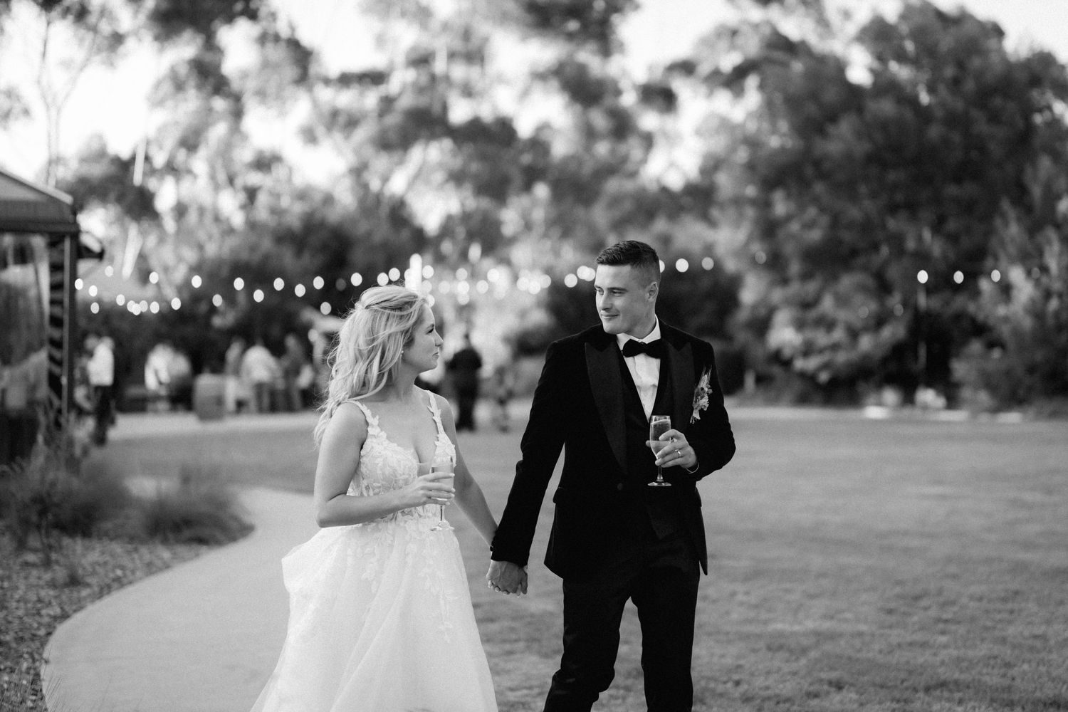 A couple in wedding attire walks together on a garden path with string lights illuminating the evening scene.
