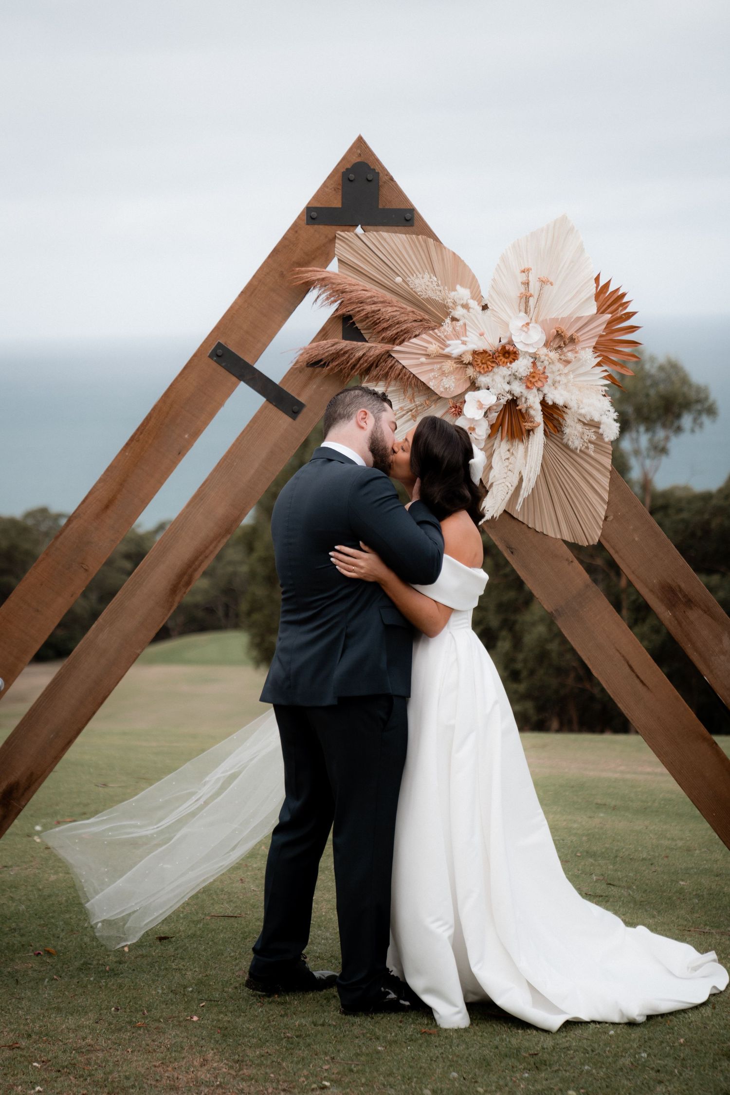A romantic wedding ceremony under a triangular wooden arch decorated with dried pampas grass arrangements on a grassy hillside.