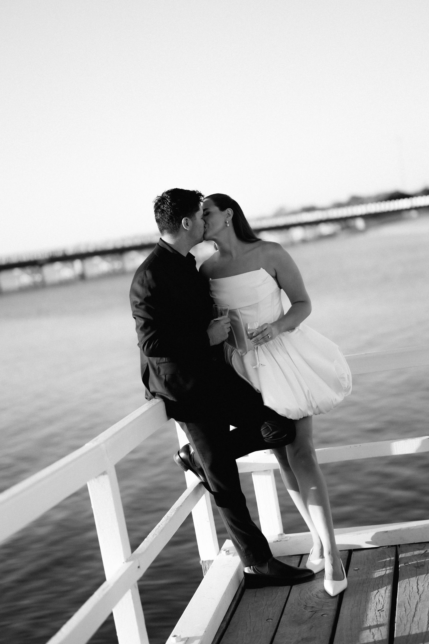 A romantic black and white photograph of a couple kissing on a white dock railing by the water.