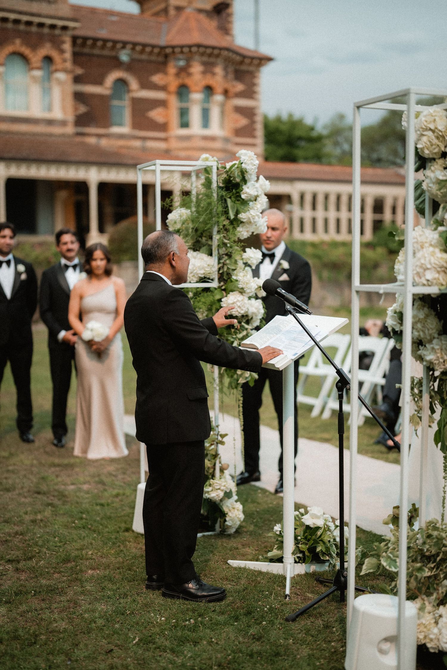 A beautiful outdoor wedding ceremony with a historical brick mansion backdrop and white floral arch decorations.