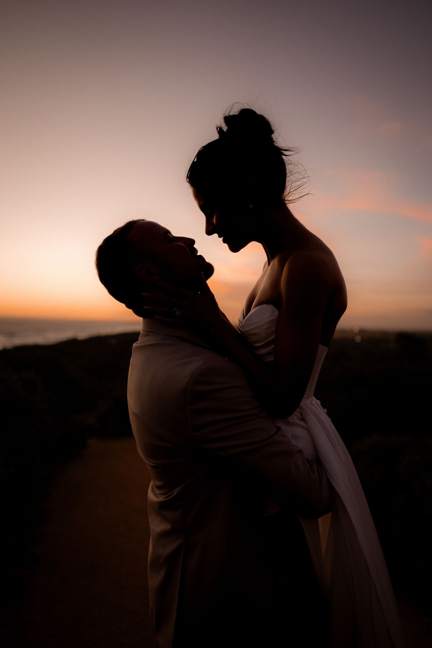 A romantic silhouette of a couple embracing at sunset, with the bride being lifted by her partner against a warm orange sky.