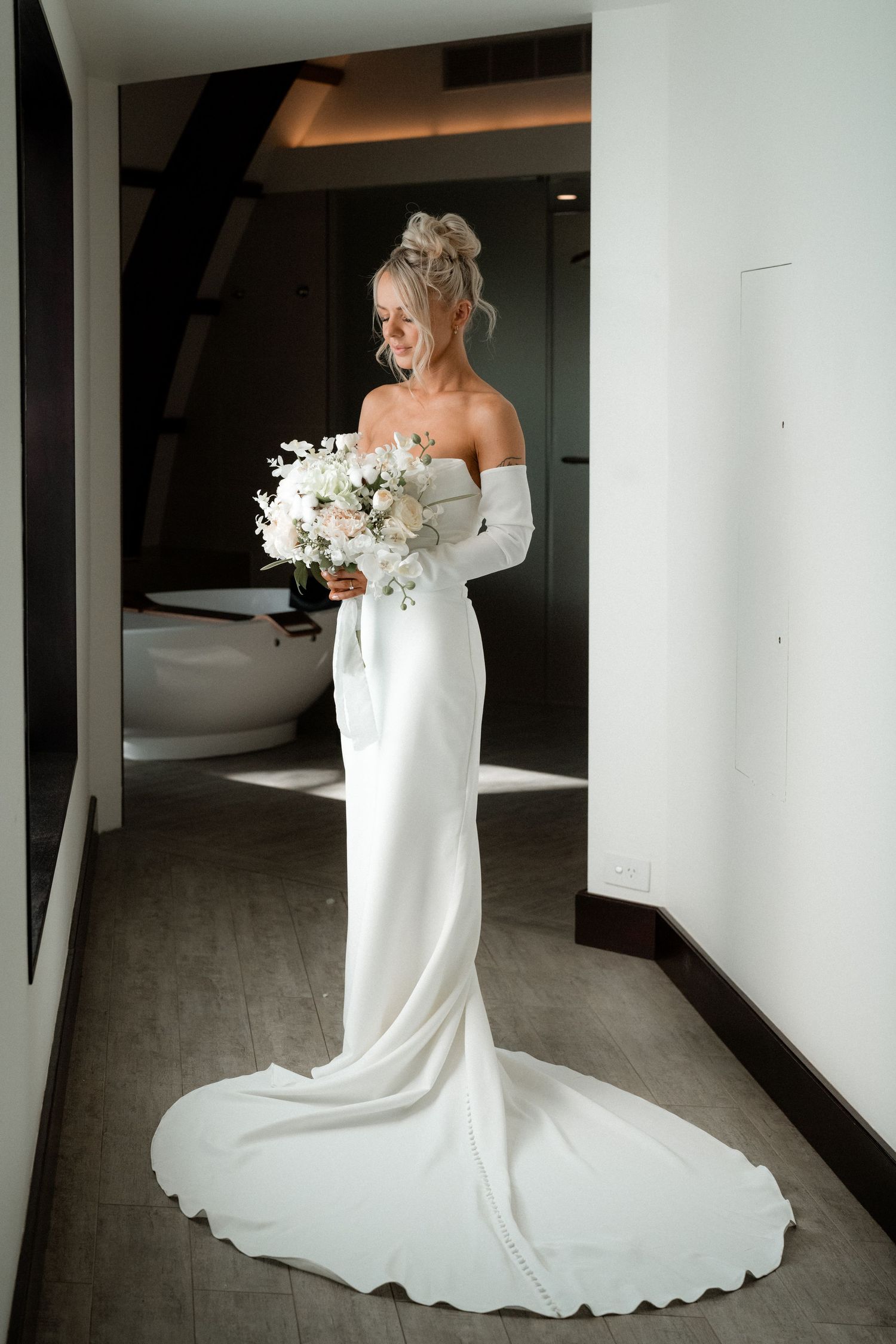 Elegant bride in a fitted white off-shoulder wedding gown holding a white floral bouquet.
