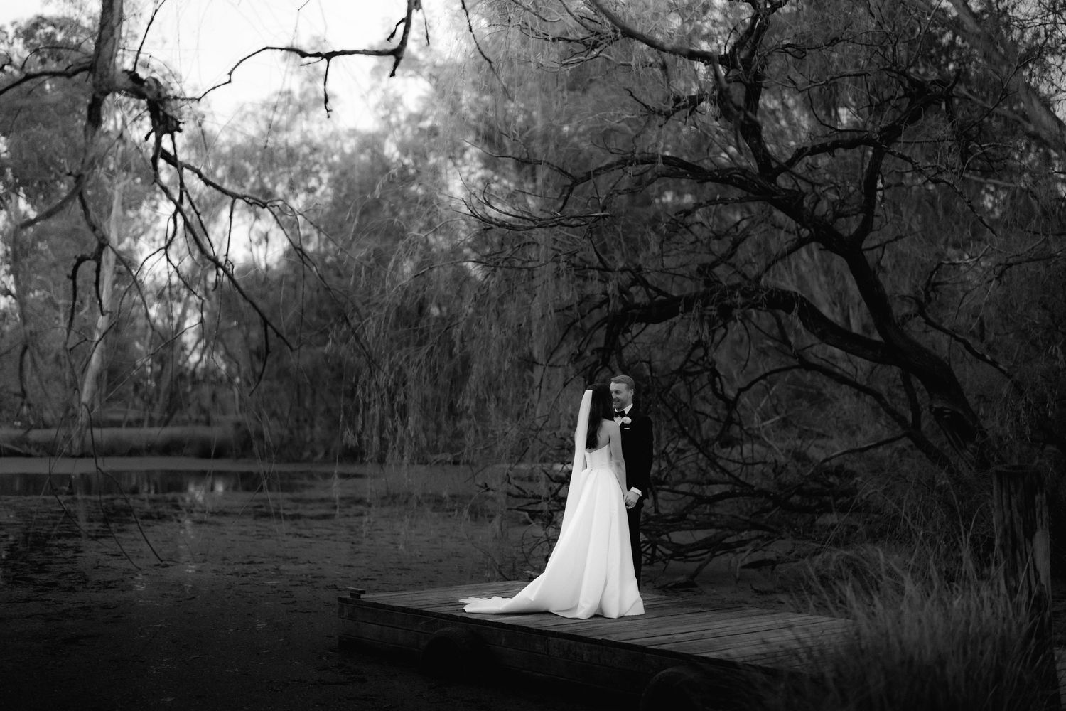 A bride in white stands on a wooden dock surrounded by moody, willow-draped scenery.