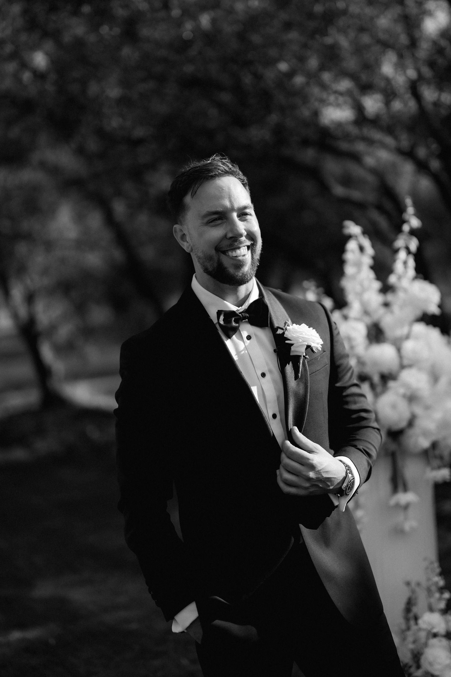 Black and white portraits of groom in tuxedo with boutonniere against natural backdrop.