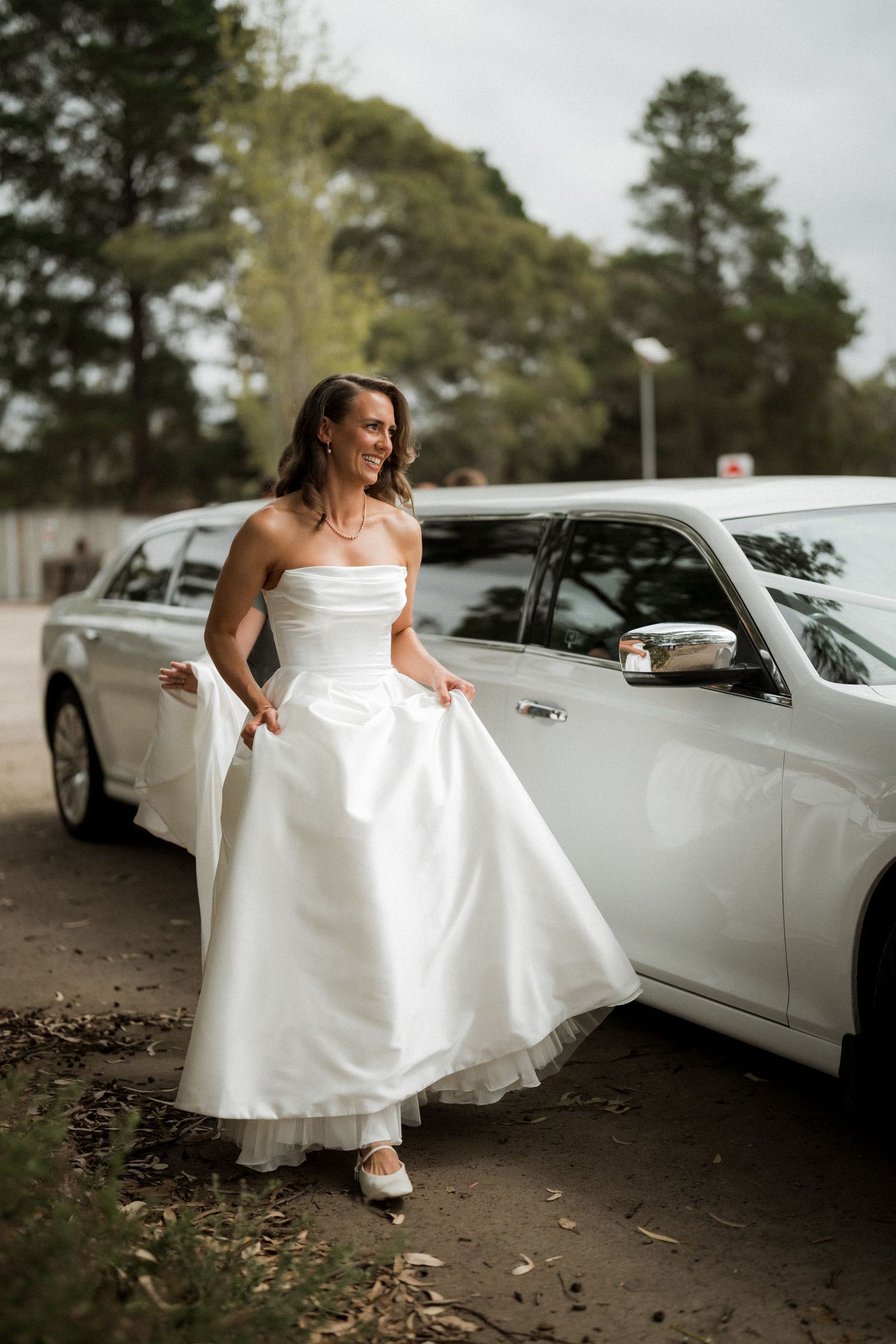 A bride in an elegant white dress poses playfully near a white limousine on her wedding day.