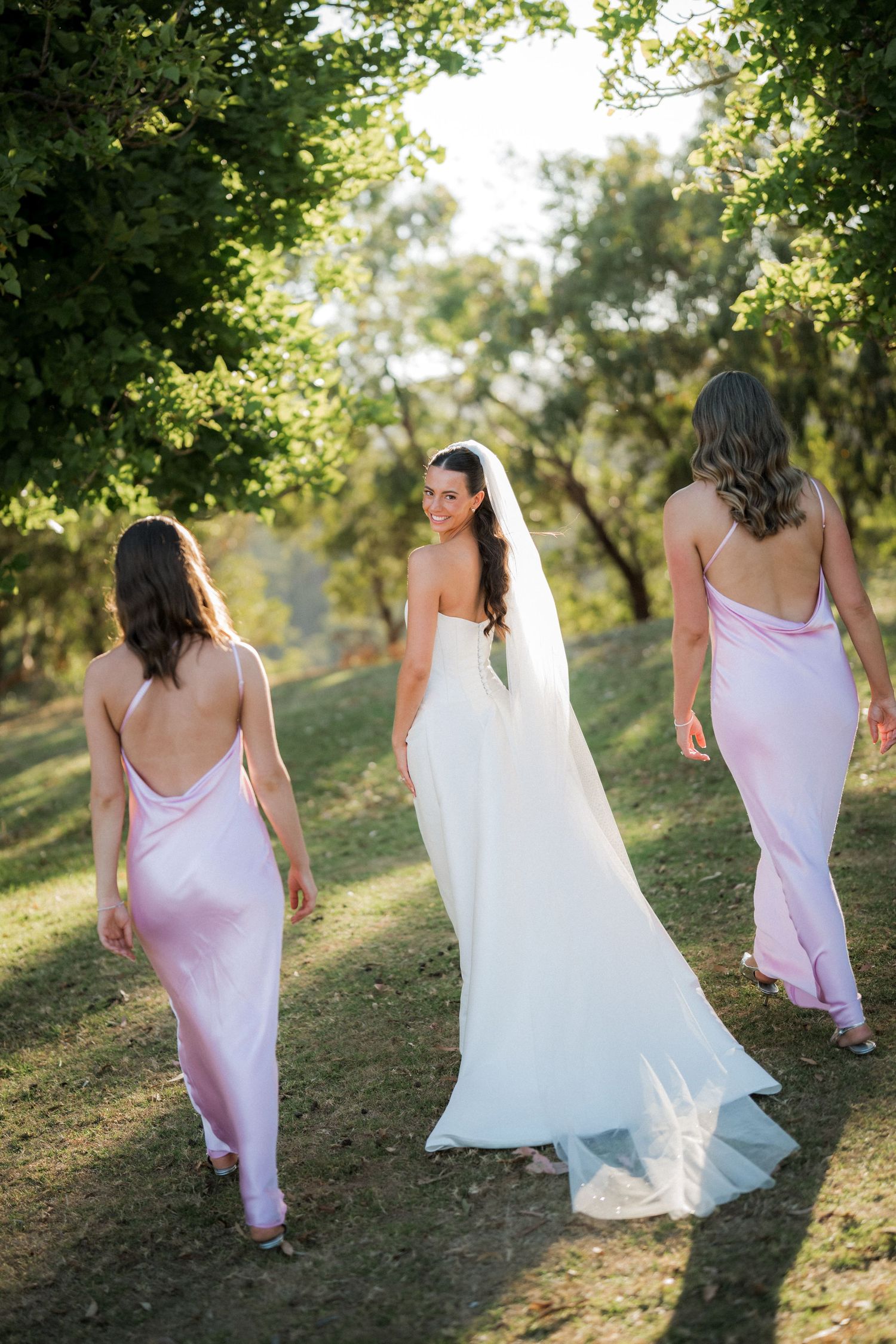 Bridesmaids in pink dresses and bride in white gown walk up grassy hill during sunset.