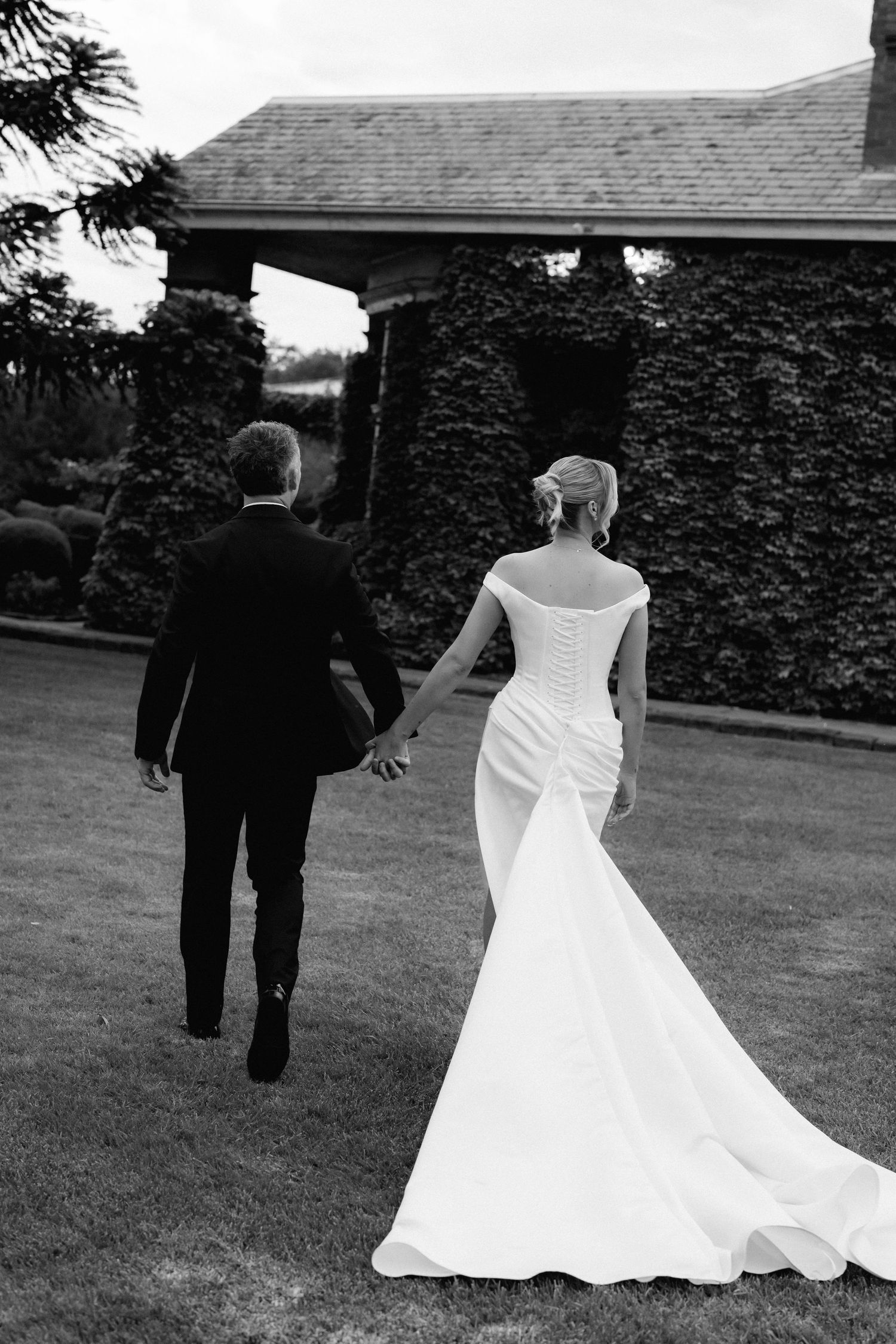 Newlyweds walking hand in hand on grass with ivy-covered building in background.