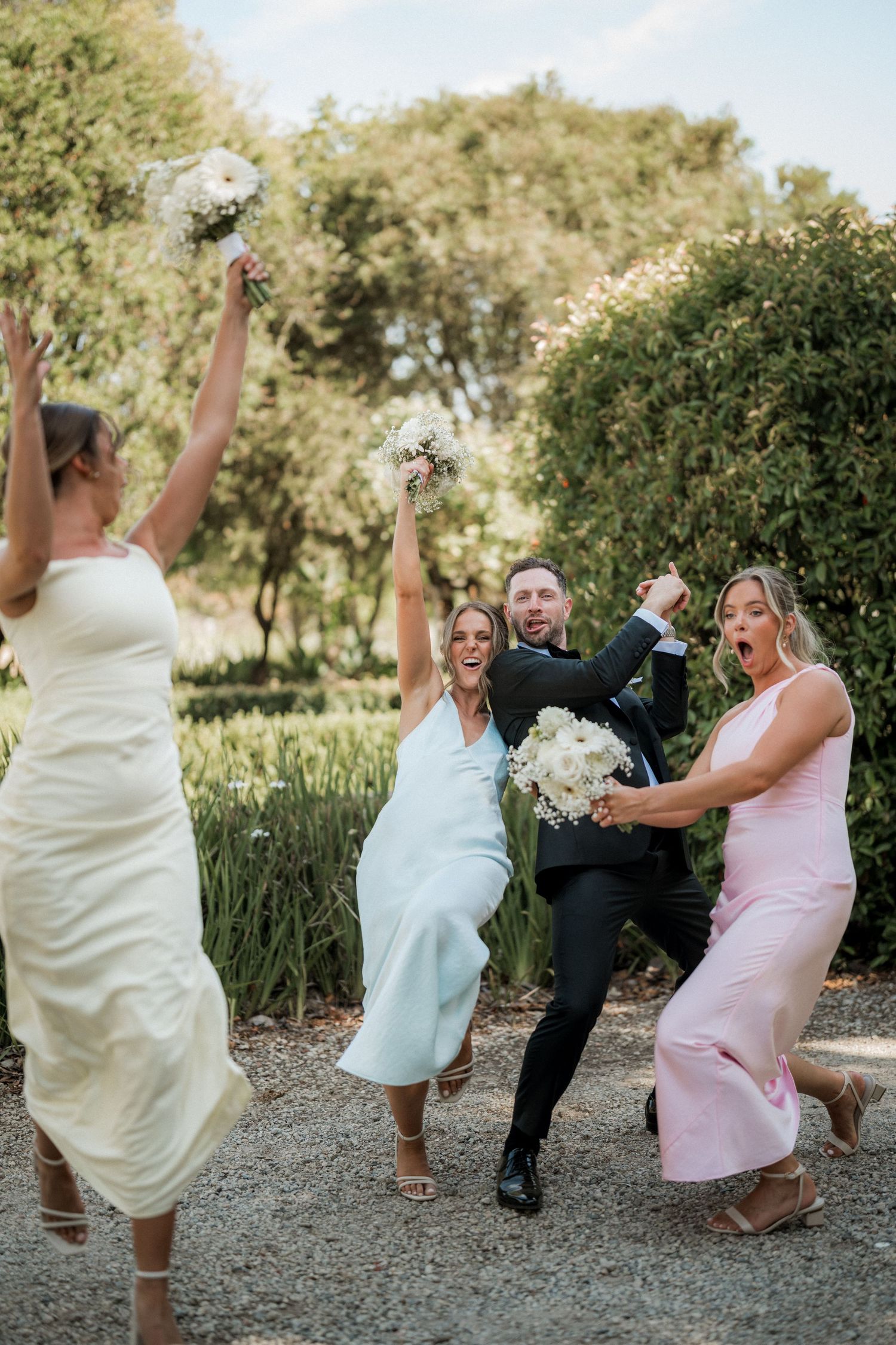 Wedding party poses playfully on a gravel path surrounded by greenery during an outdoor photoshoot.
