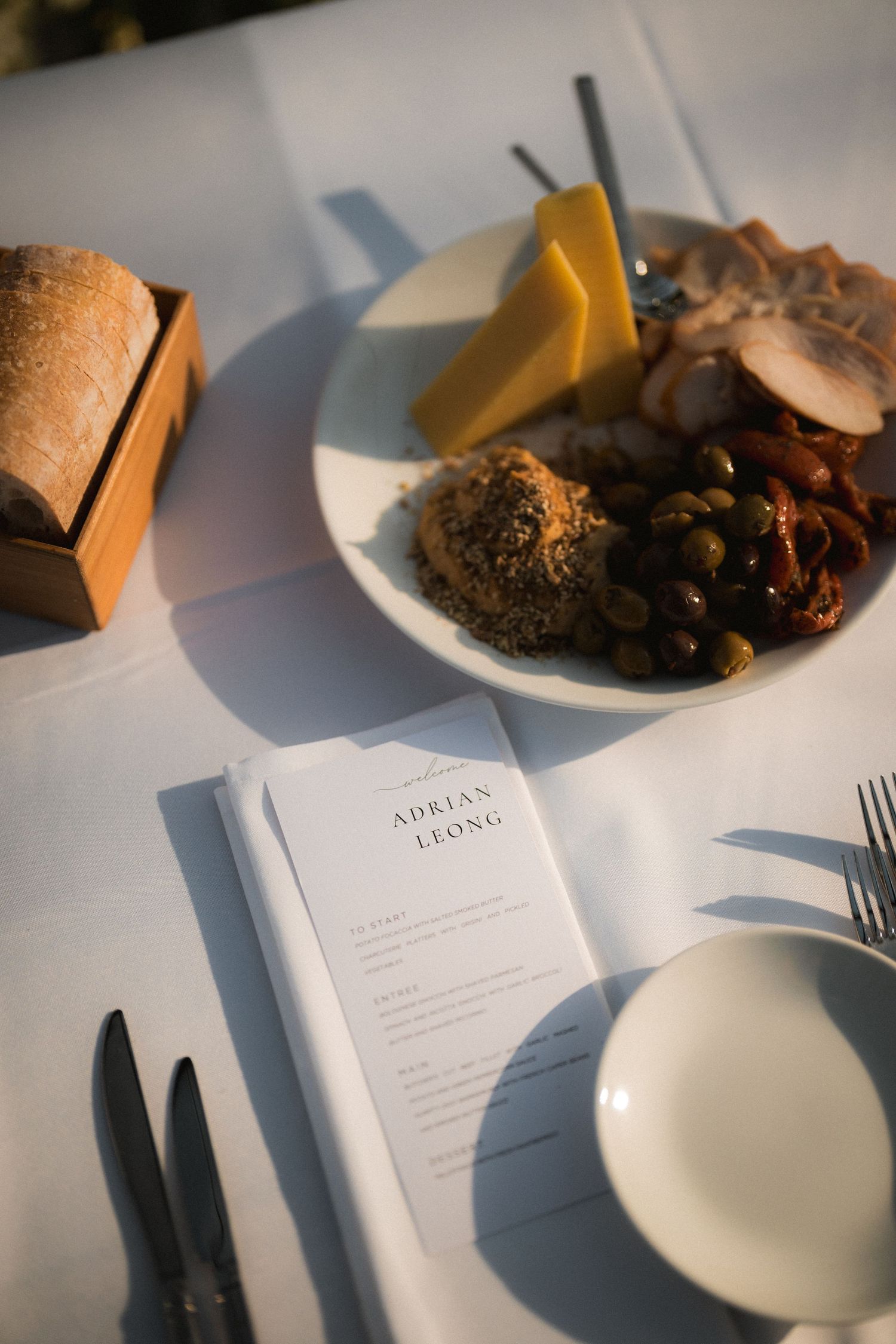 Wedding table setting with a charcuterie plate of cheese, olives and sliced meats beside a bread box and a printed menu card.