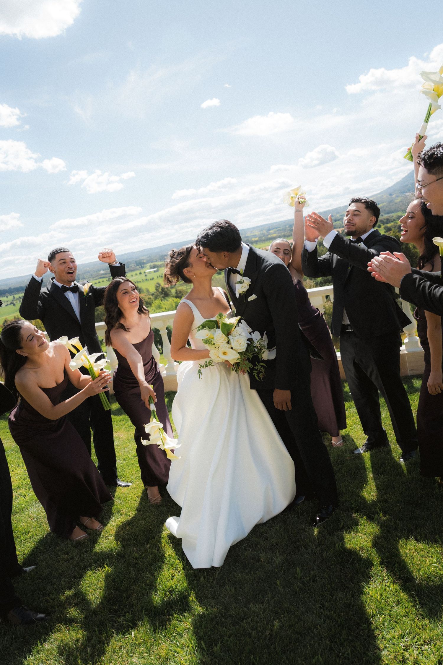 Bride in a white gown kisses the groom as cheering bridesmaids and groomsmen celebrate on a sunny hilltop lawn at an outdoor wedding.