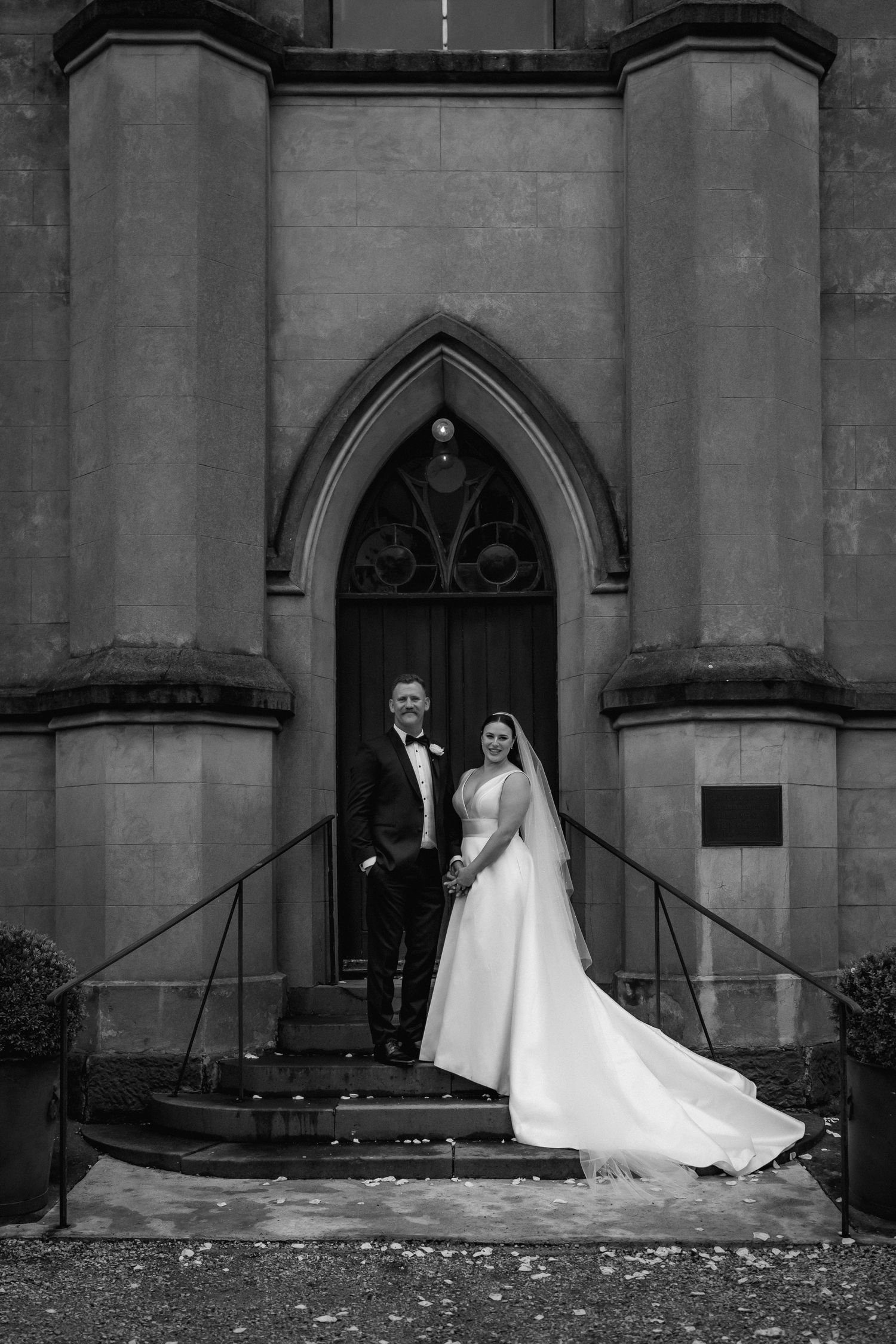 A black and white photo of a couple posing in formal attire at an elegant gothic church doorway.