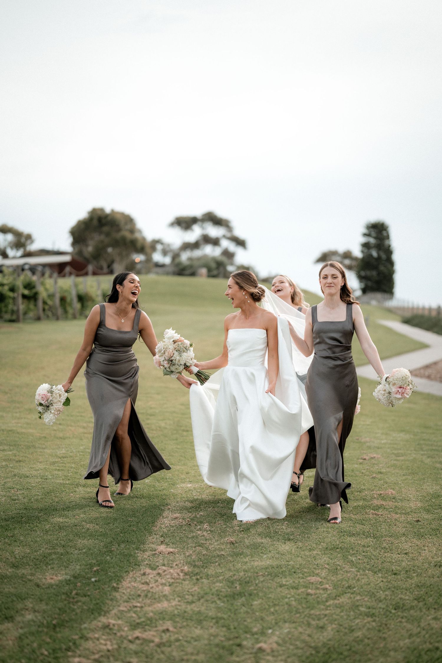 A wedding party in grey bridesmaid dresses and white bridal gown runs joyfully across a grassy hill while holding bouquets.