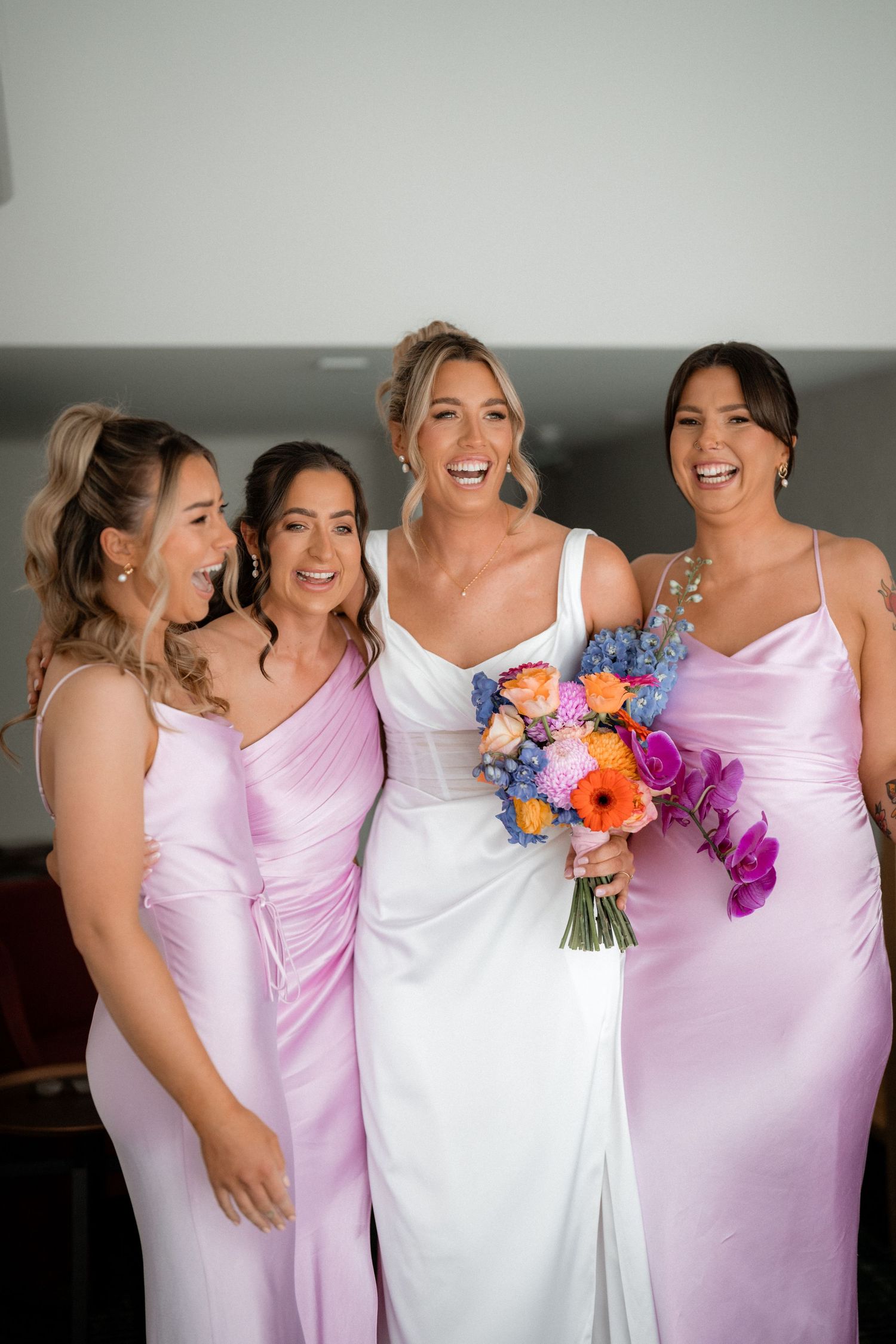 A bridal party wearing pink satin dresses pose together with a vibrant colorful bouquet of flowers.