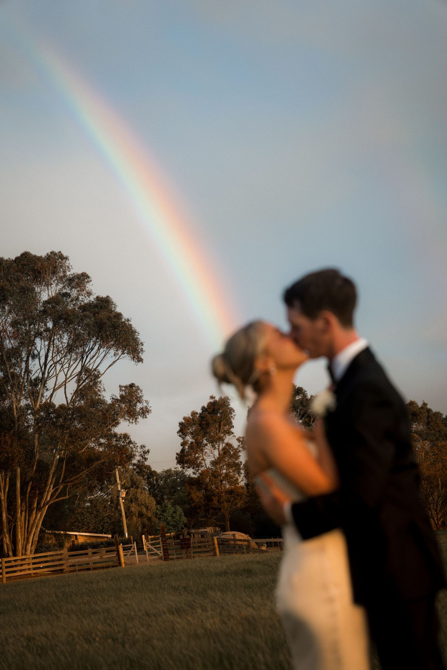 Wedding couple embraces against a dramatic sky with a rainbow in the background at sunset.