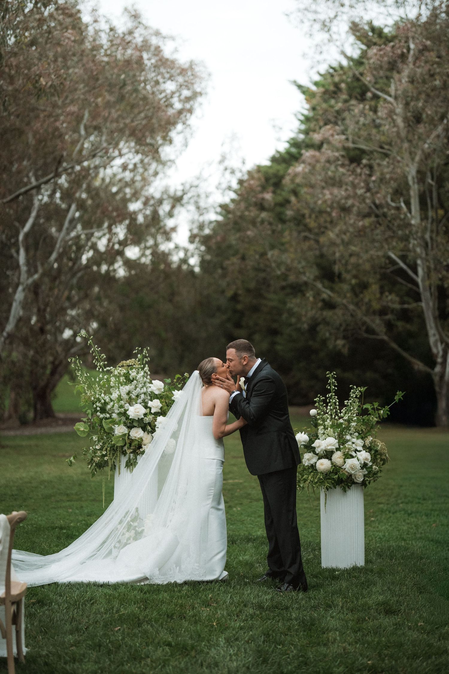 Outdoor wedding ceremony setup with white floral arrangements and tall trees in the background.