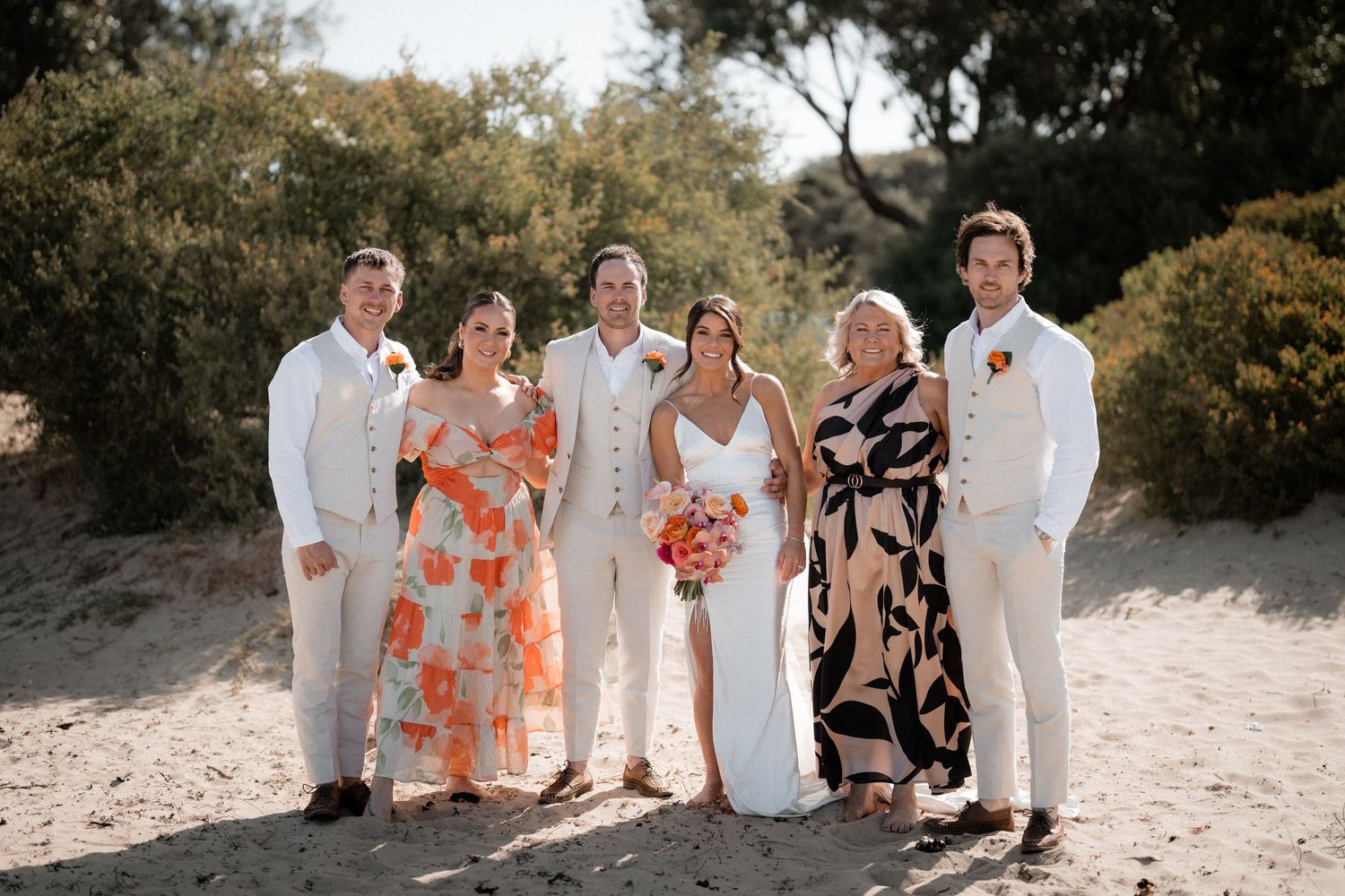 Wedding party in white and coral attire poses together on a sandy beach path.