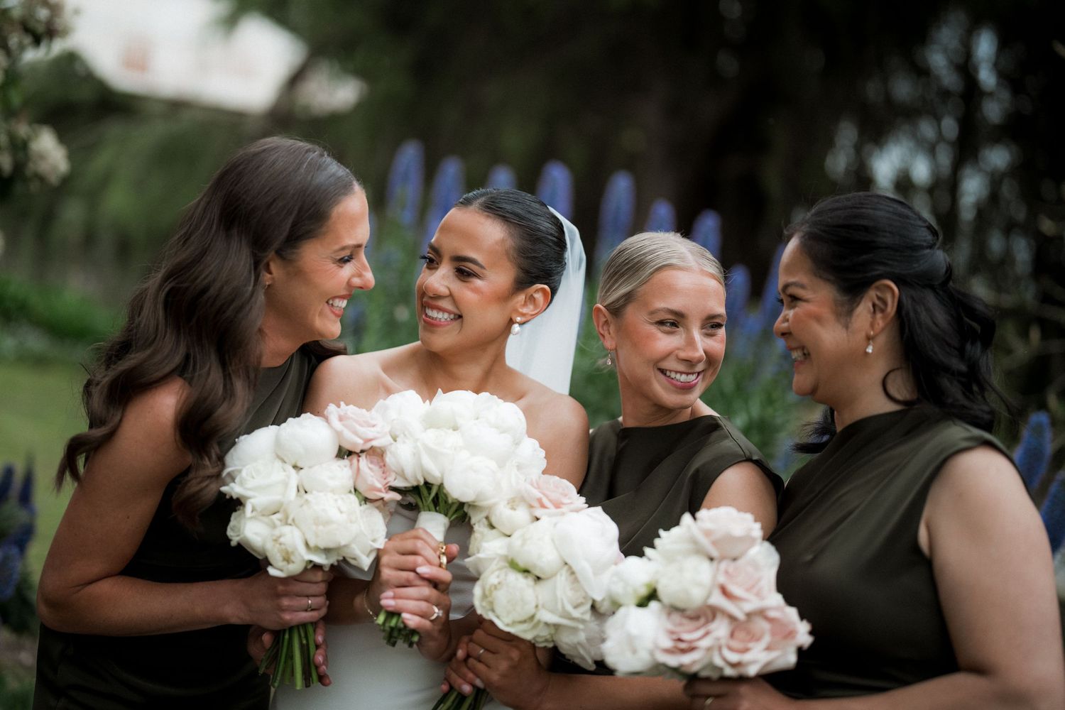 Bridesmaids in elegant dark dresses share joyful laughter while holding beautiful white bouquets at an outdoor wedding.