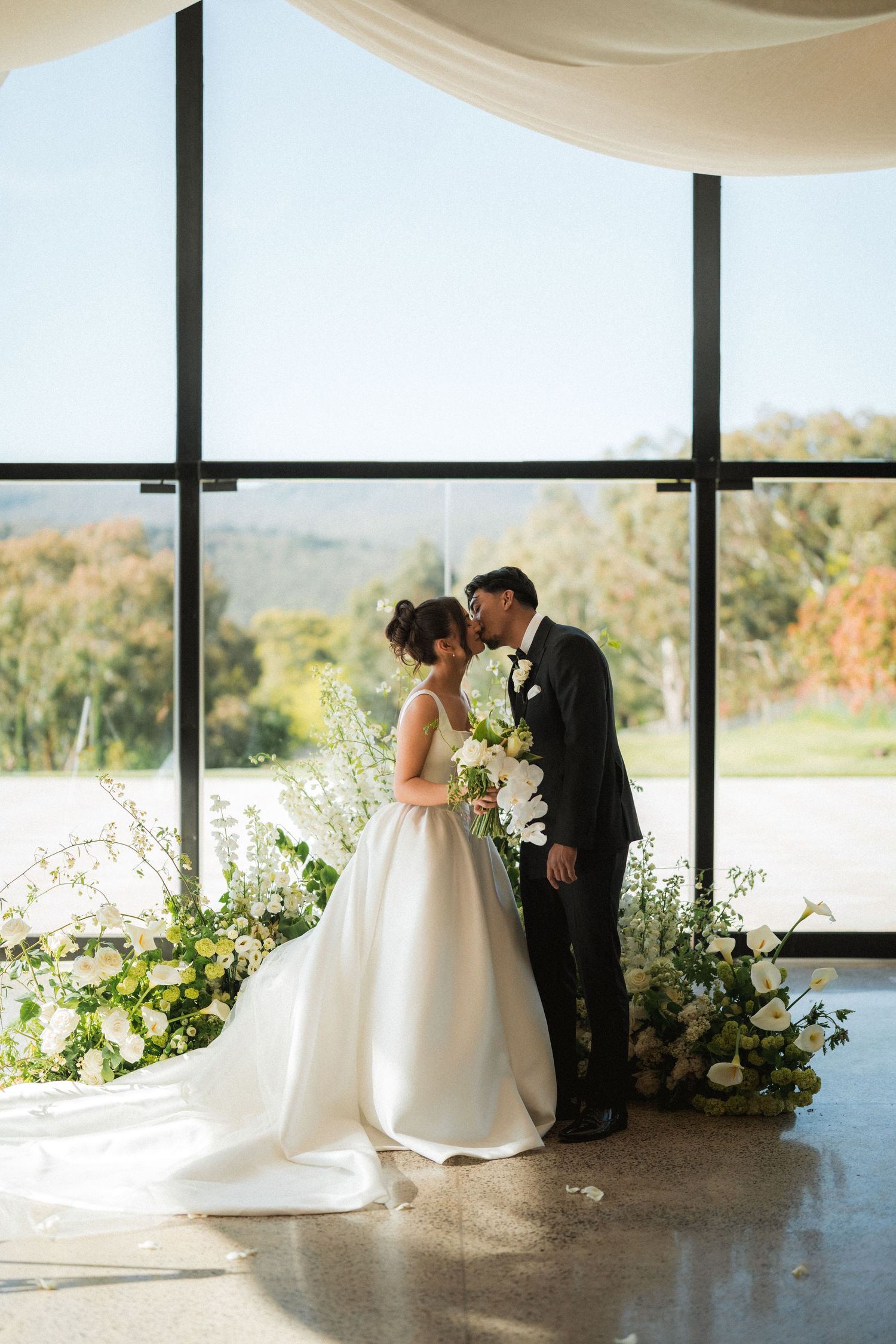Bride and groom share a kiss surrounded by white floral arrangements in front of large windows with scenic garden views.