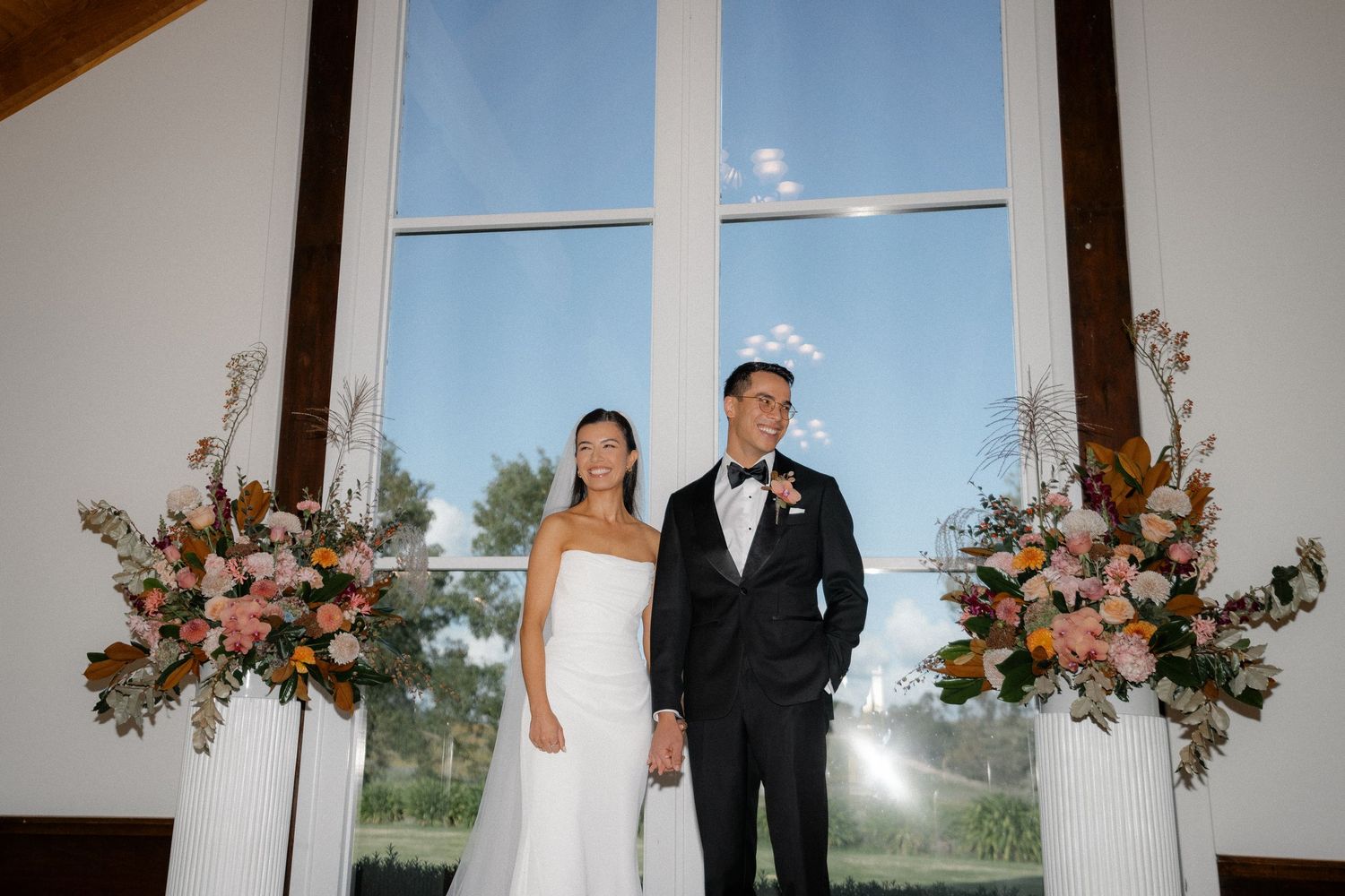 Bride and groom stand at chapel altar flanked by pink and white floral arrangements.