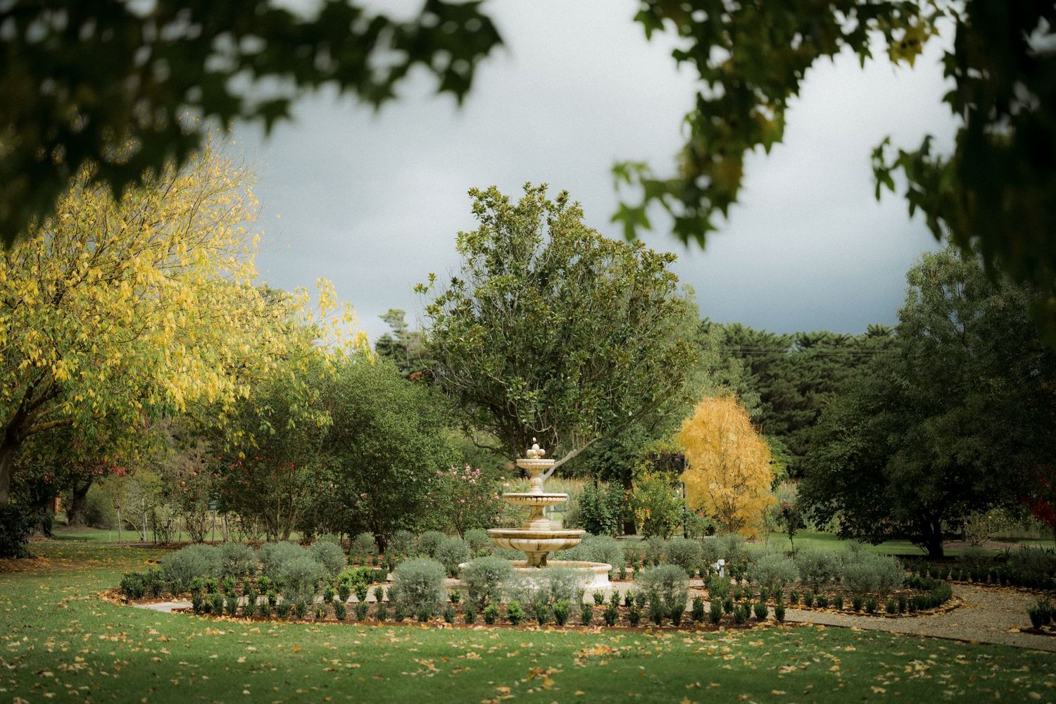 Serene garden fountain surrounded by lush trees and manicured hedges under a cloudy sky.