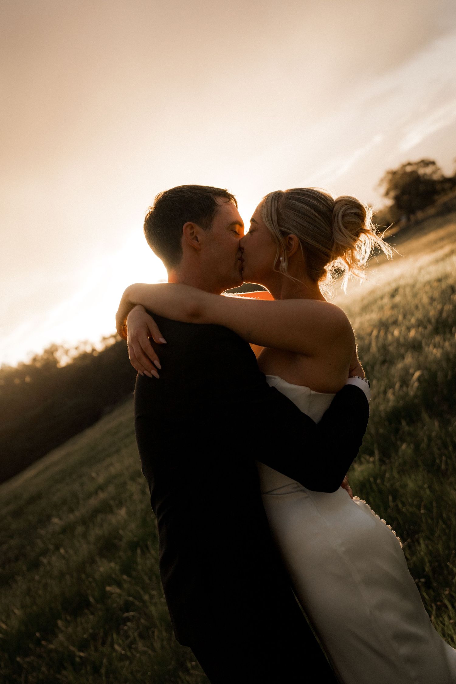 Newlyweds share an intimate moment at sunset in a grassy field.