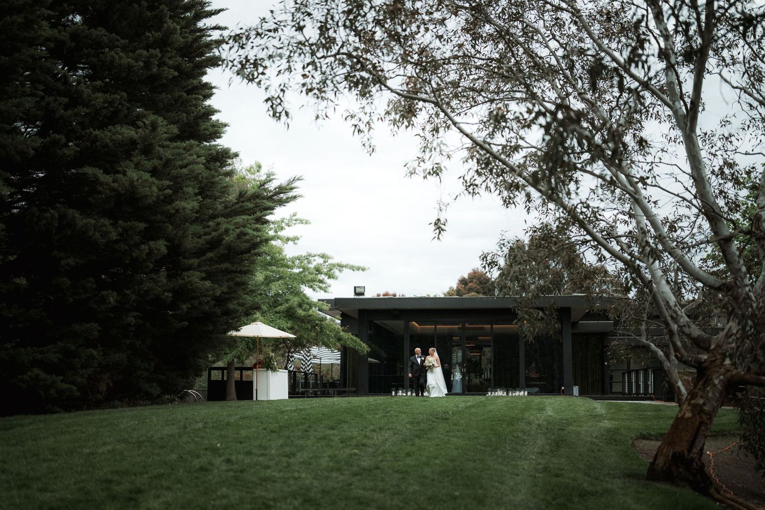 Modern wedding venue with lush green lawn and contemporary architecture surrounded by tall trees.