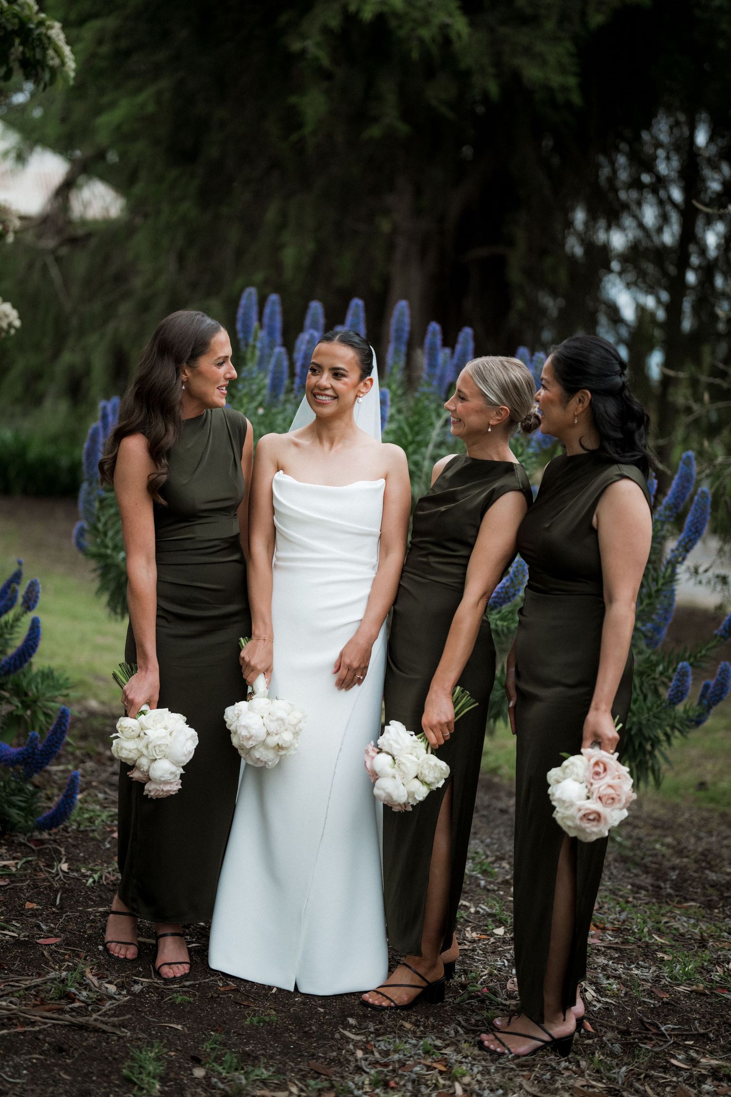 Wedding party poses outdoors with bridesmaids in brown dresses and white bouquets against a natural backdrop.