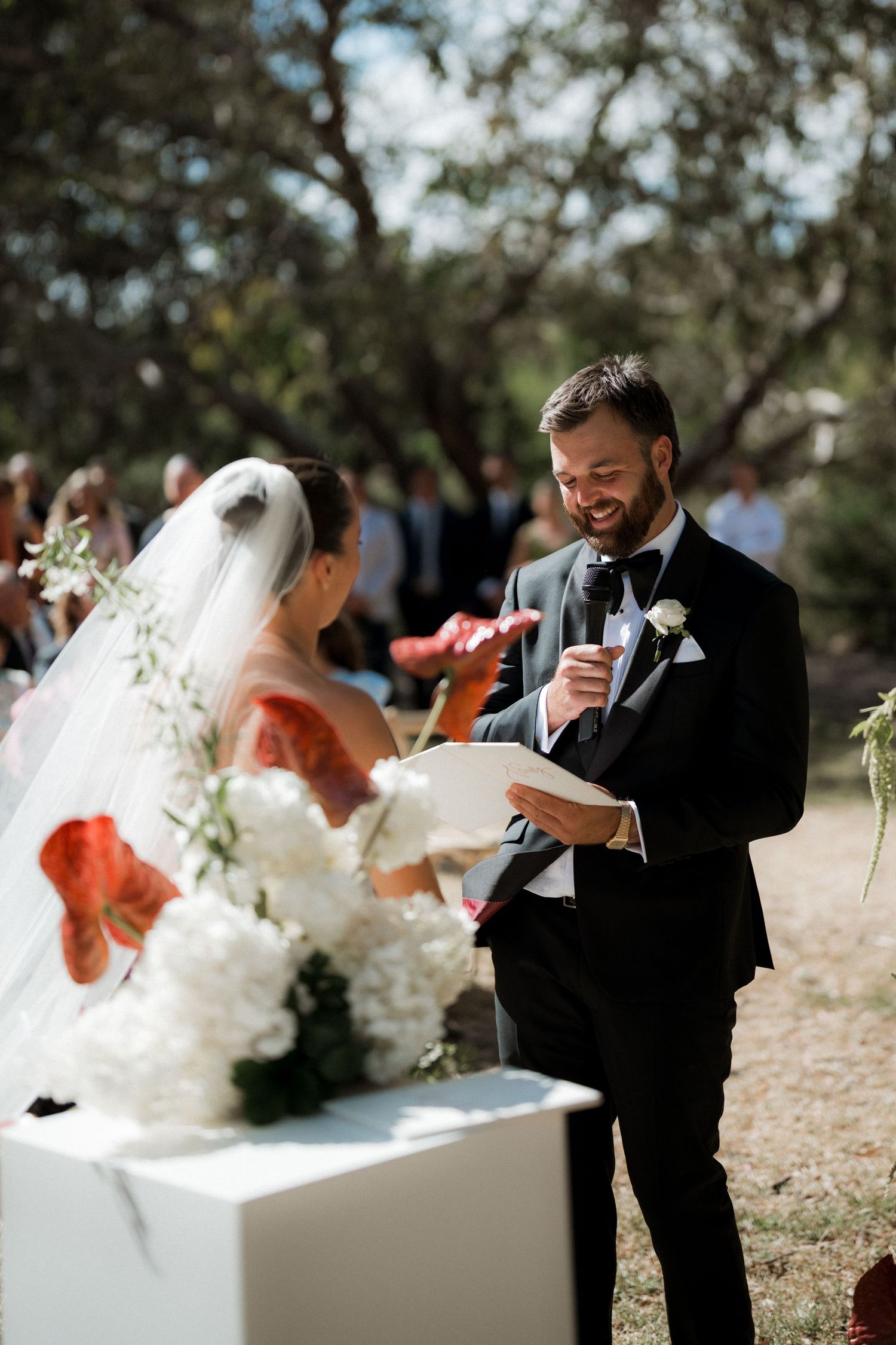 Groom in black tuxedo smiling while reading vows to bride in white dress and veil at outdoor wedding ceremony.