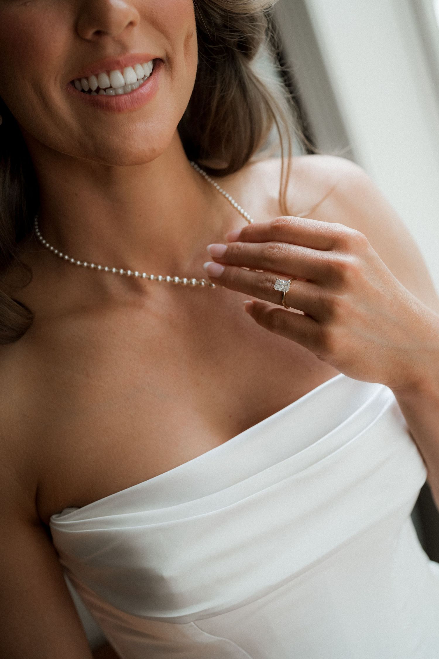 Close-up details of a white off-shoulder wedding dress with delicate pearl necklace and engagement ring.