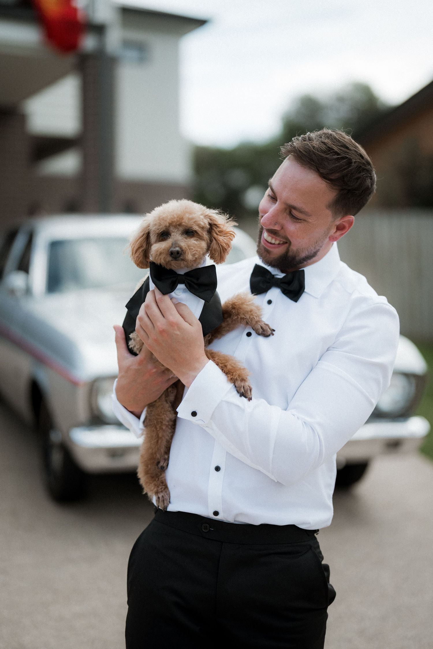 A groom in white tuxedo holding an adorable small dog wearing a matching black bowtie.