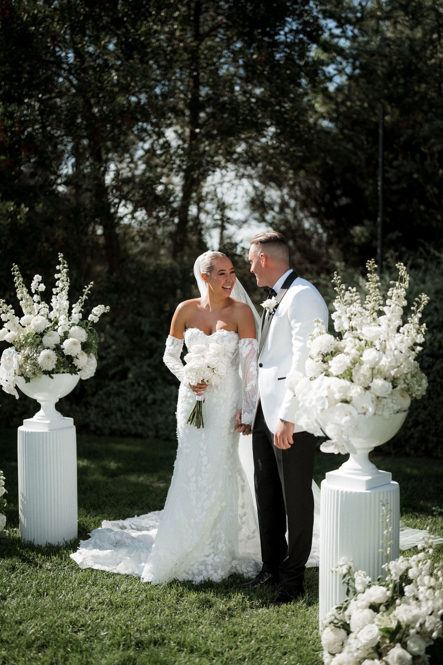 Elegant outdoor wedding ceremony setup with white floral arrangements and decorative urns on pedestals.