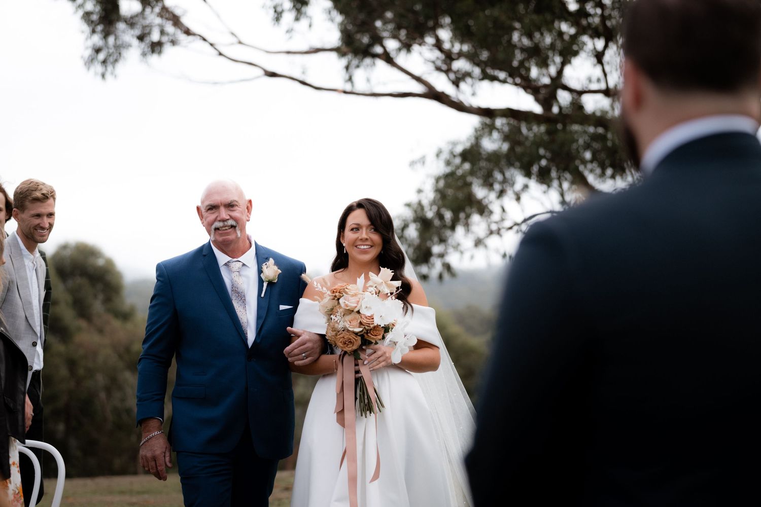 A bride in a white dress holds a bouquet while walking outside with someone in a navy suit on a cloudy day.