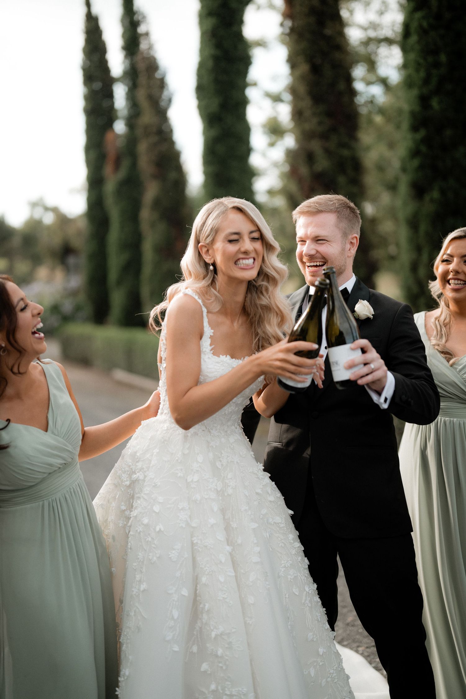 Wedding party sharing laughs and drinks amongst tall cypress trees.