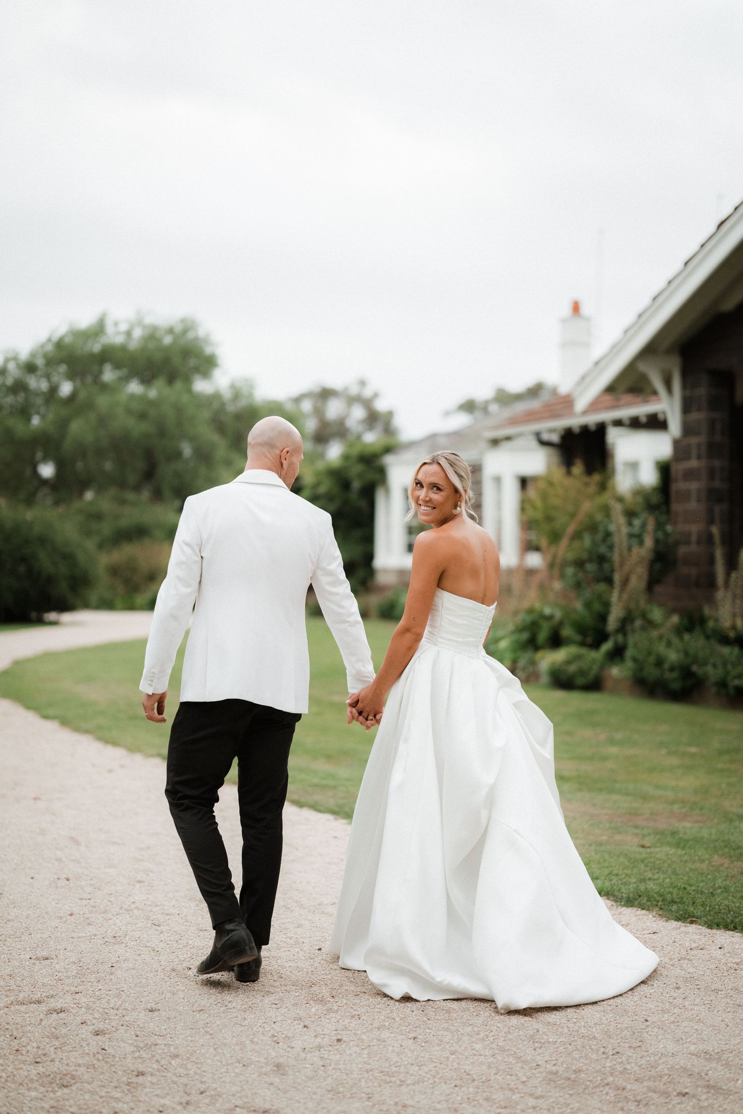 Couple in formal wedding attire walking down garden path holding hands.