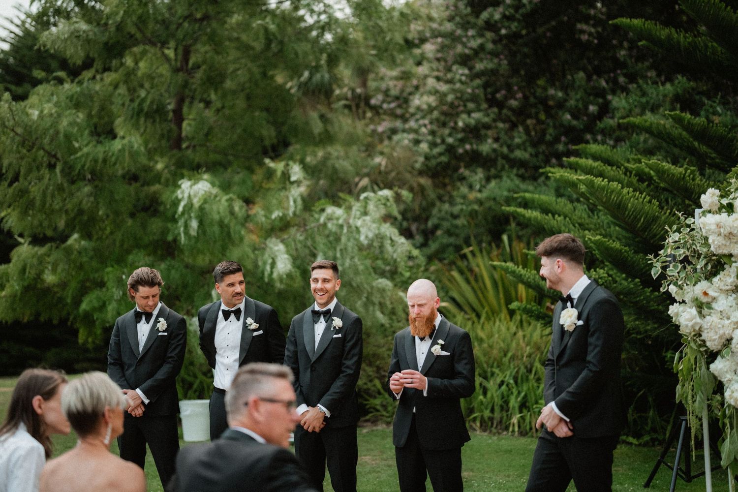 Wedding party gathered at an outdoor garden ceremony with lush greenery and white floral arrangements.