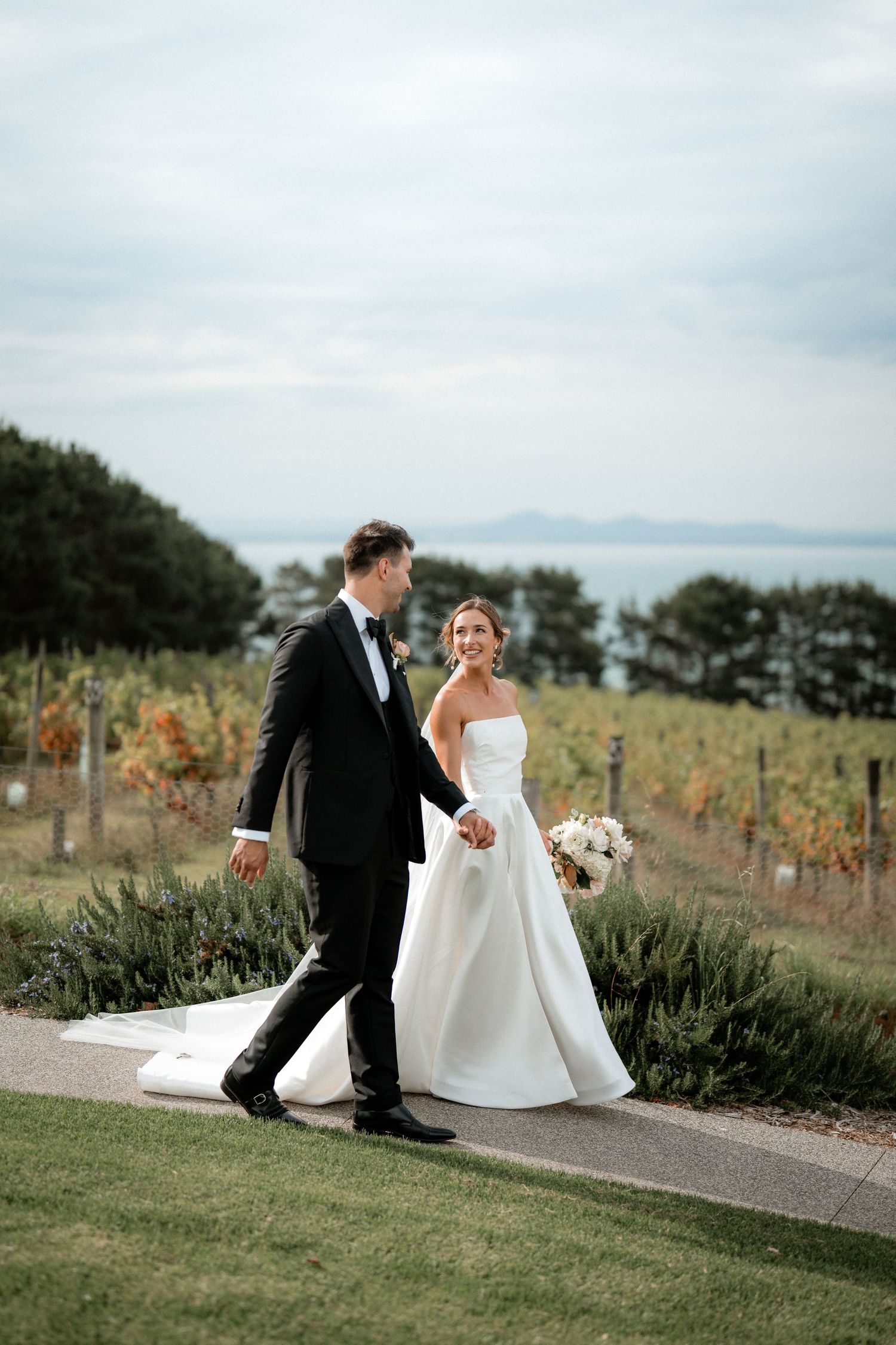 A wedding couple walks together on a path overlooking a vineyard and water view on their special day.