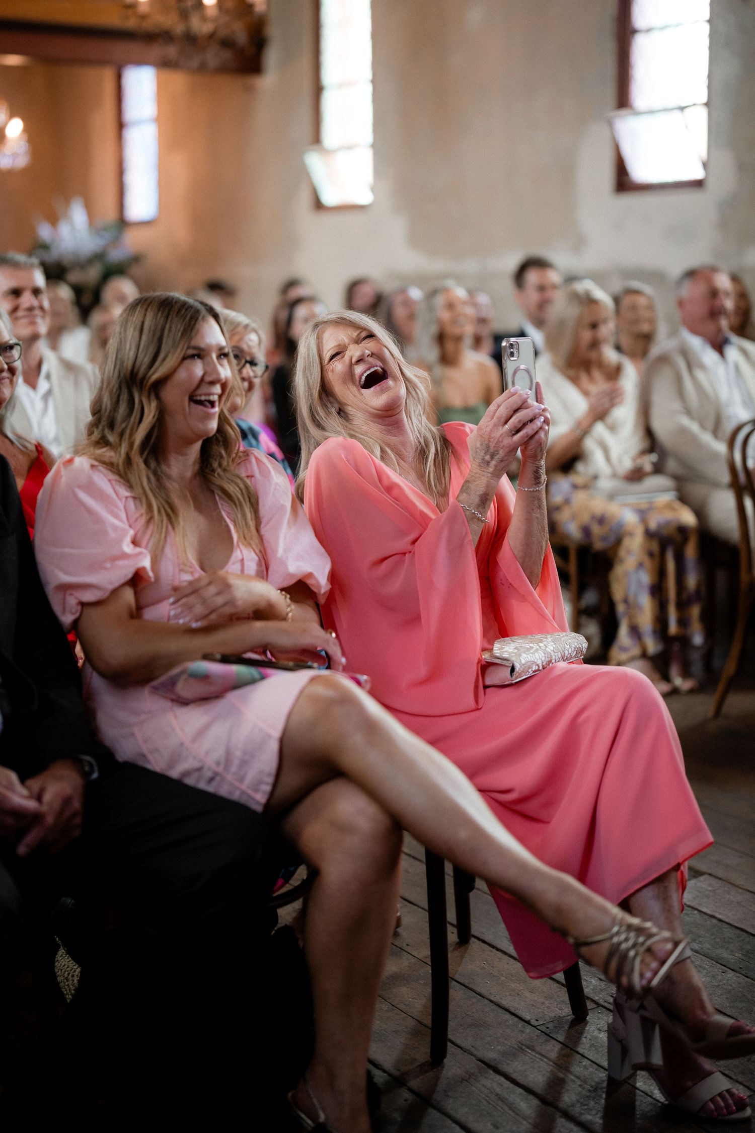 Two women in pink dresses share a laugh together while sitting in audience at an indoor event gathering.