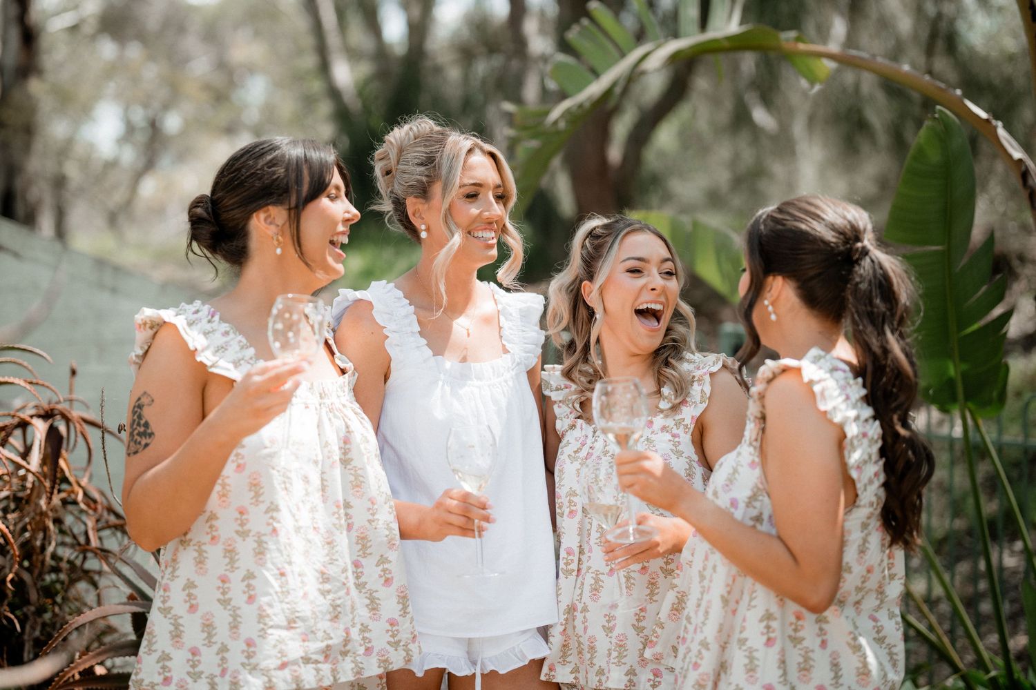 Bridesmaids in white patterned dresses laughing and having drinks outdoors.