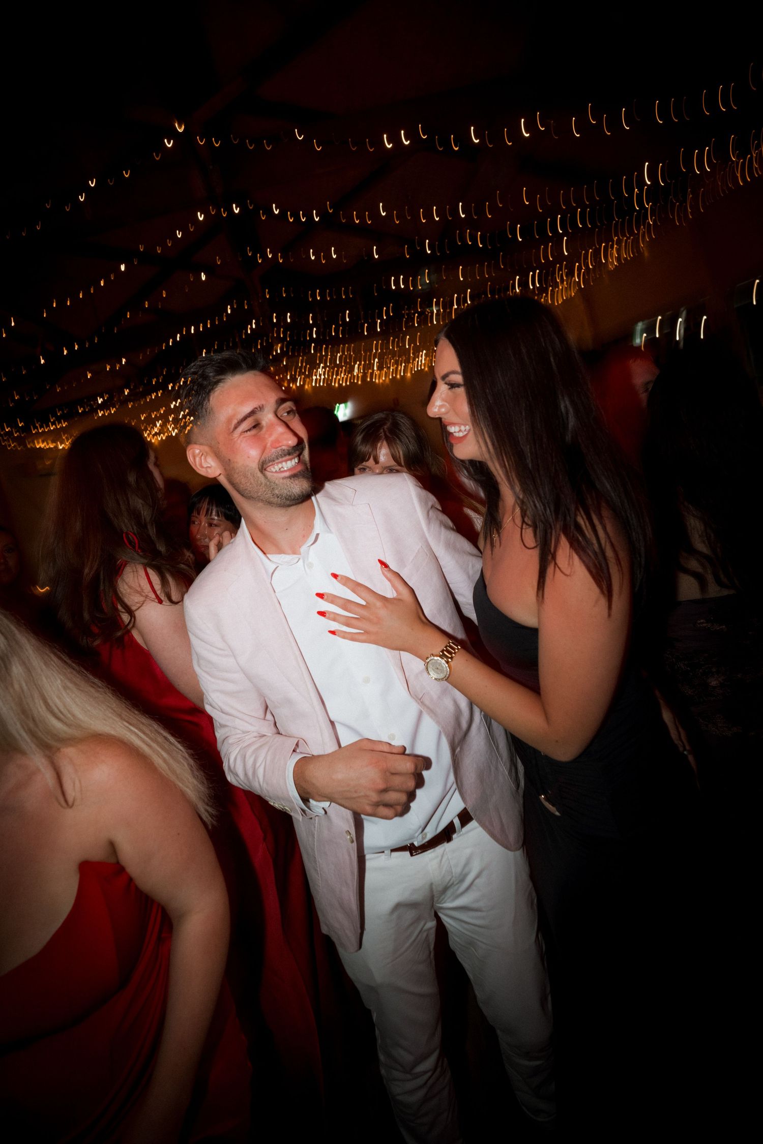 Guests dancing and celebrating at a dimly lit party venue with string lights overhead.