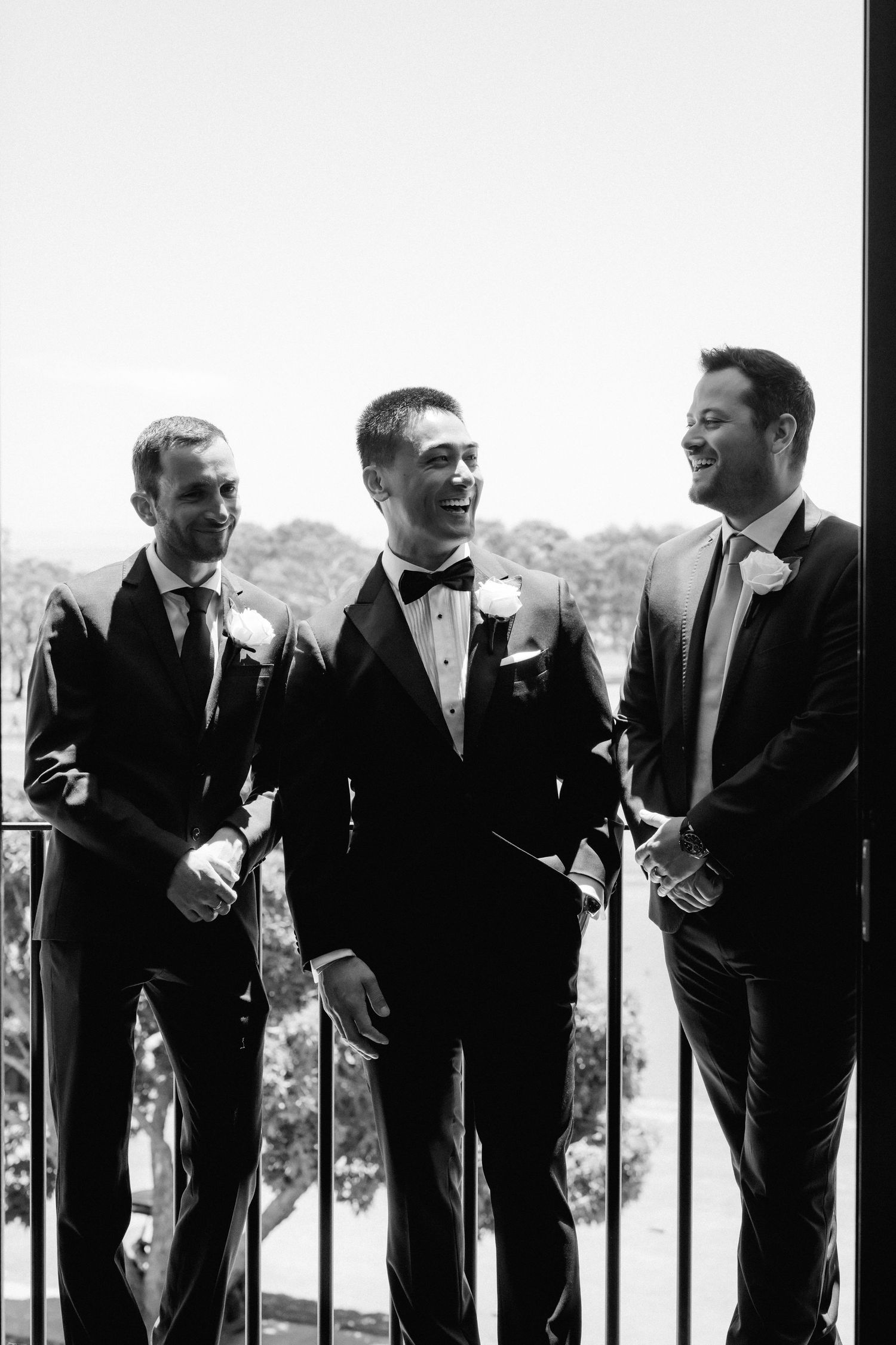Three groomsmen in formal attire share a candid laughing moment on a balcony in black and white.