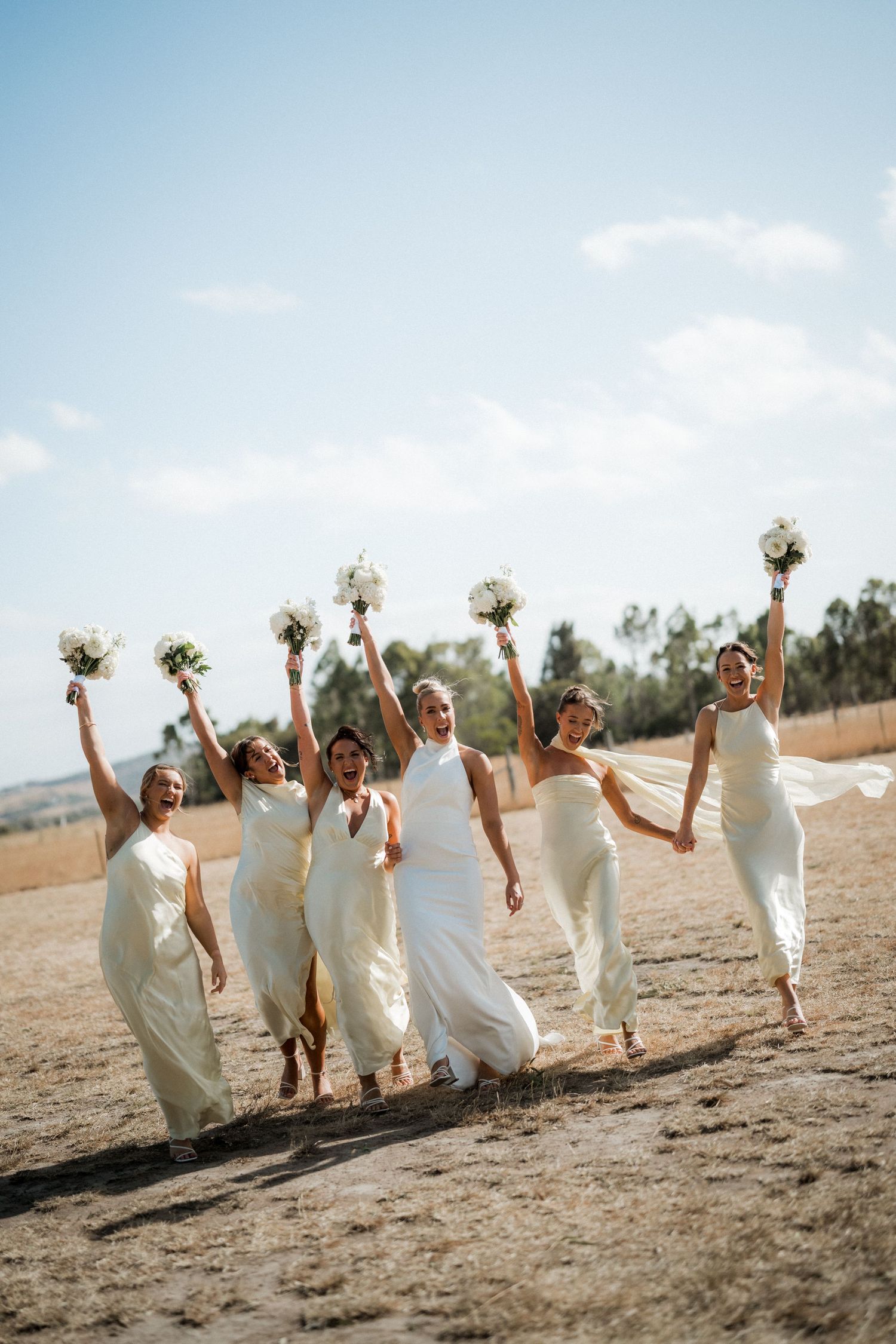 Bridesmaids in white dresses run and jump together on a sunny beach holding bouquets.