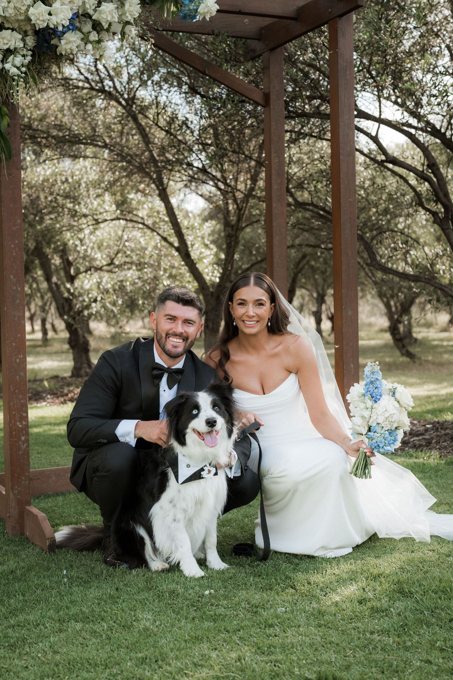Wedding couple poses with their black and white dog under wooden pergola in garden setting.