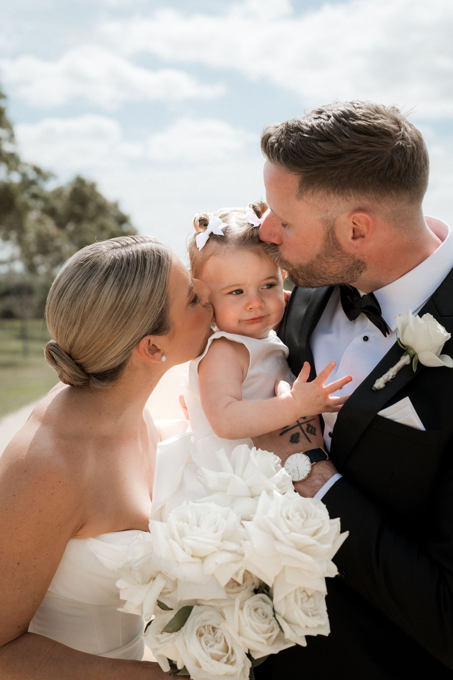 Sweet family moment with bride and groom kissing their small child surrounded by white roses.