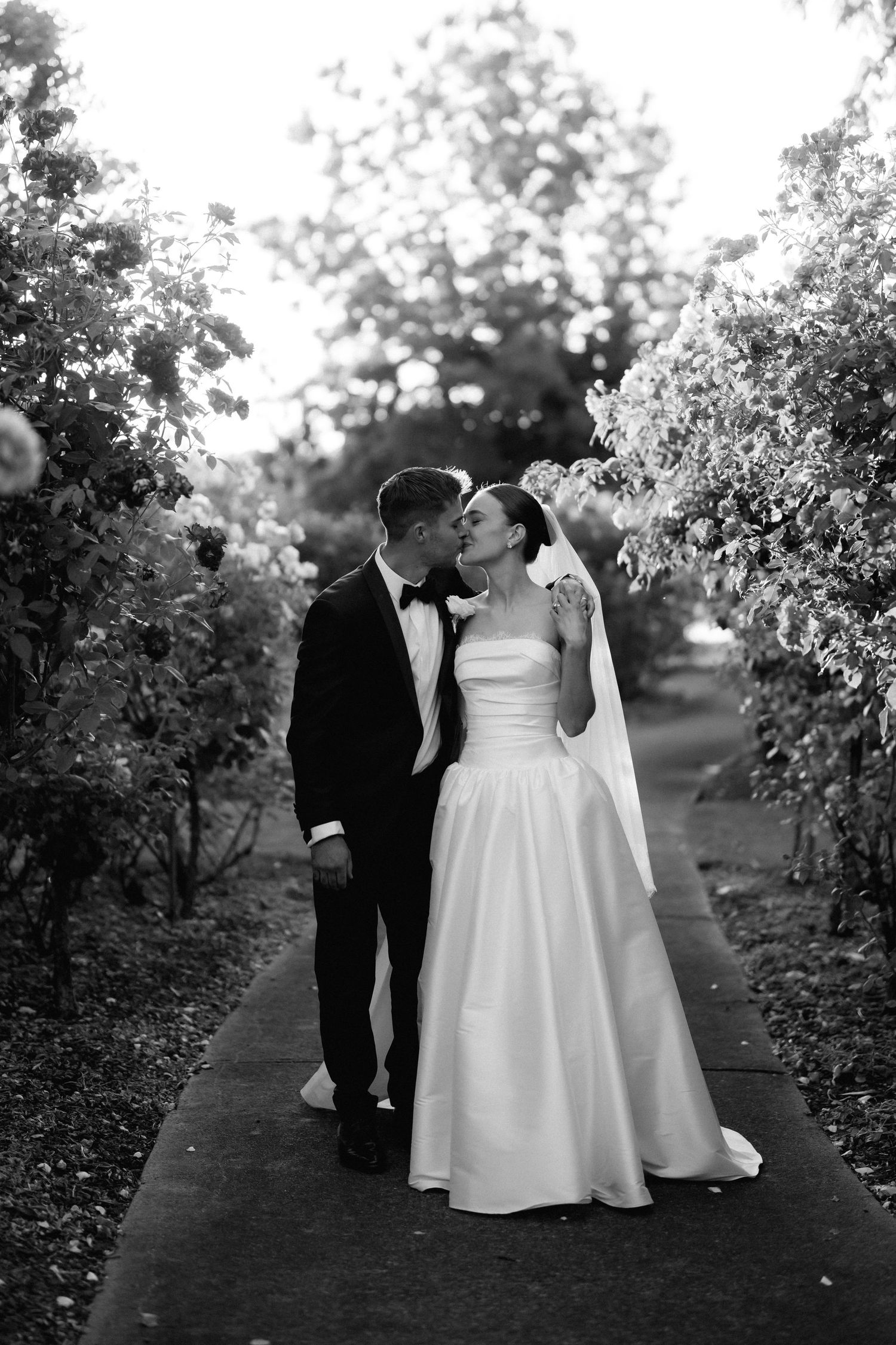 A couple walks through an orchard path during their wedding photoshoot in black and white.